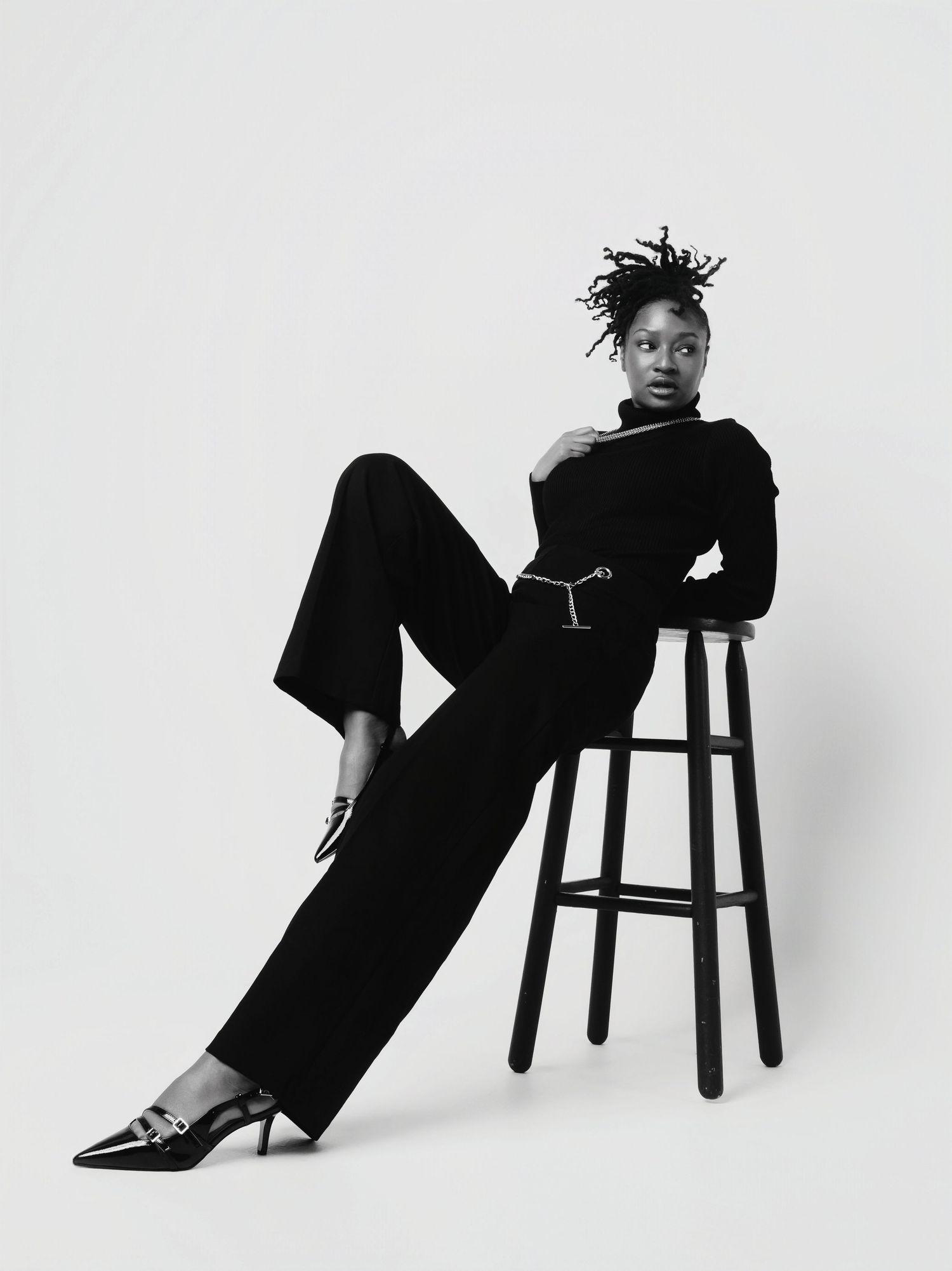 Black and white fashion photograph showing a person in all-black attire posing elegantly on a wooden barstool.