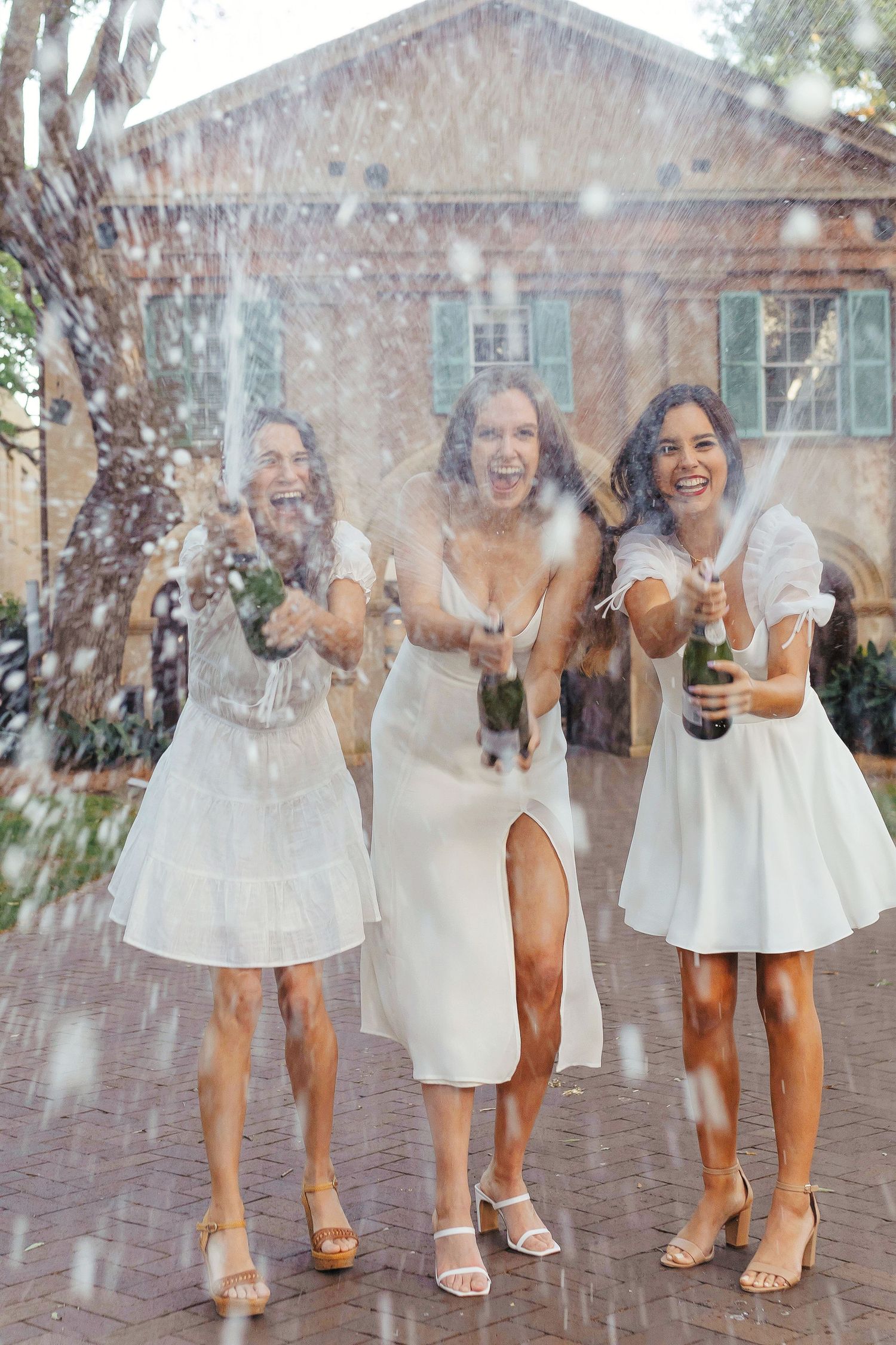 Three women in white dresses stand together laughing and celebrating as confetti falls around them on a brick patio.