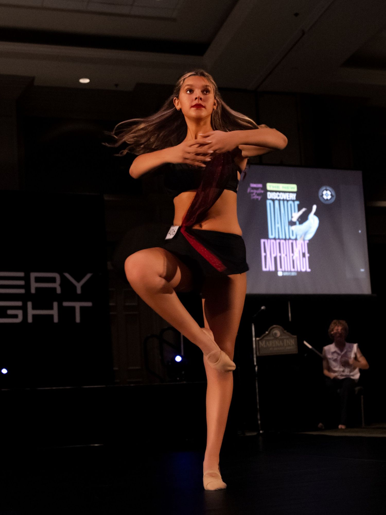 A dancer performs a high kick sequence on stage during a dance performance with dramatic lighting and projection screens.