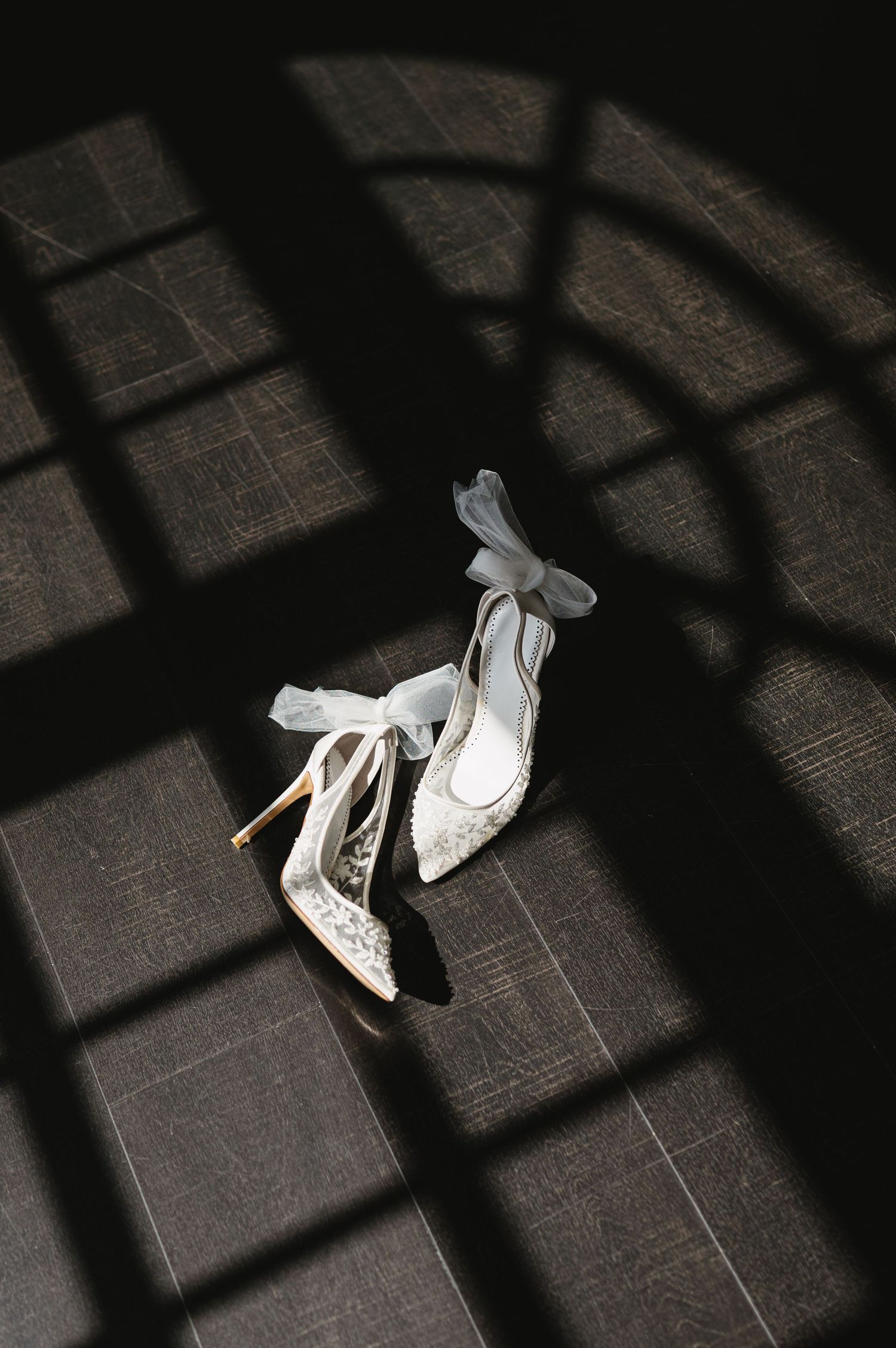 Elegant white bridal heels with bow details cast dramatic shadows on dark tiled floor in artistic composition.