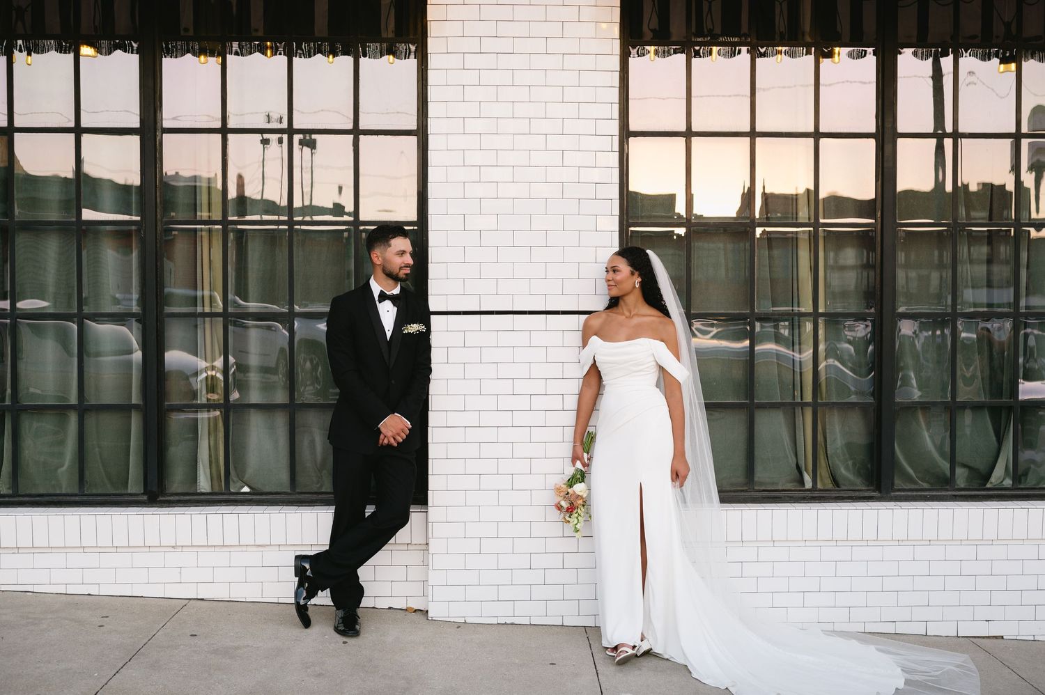 A bride in a white off-shoulder gown and groom in black tuxedo pose against a white brick wall with large windows.