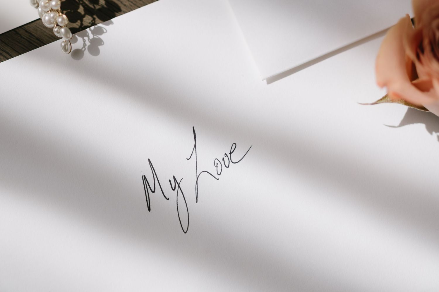 A hand holding a pen writes 'My Love' in elegant black cursive handwriting on white paper with small flowers visible.
