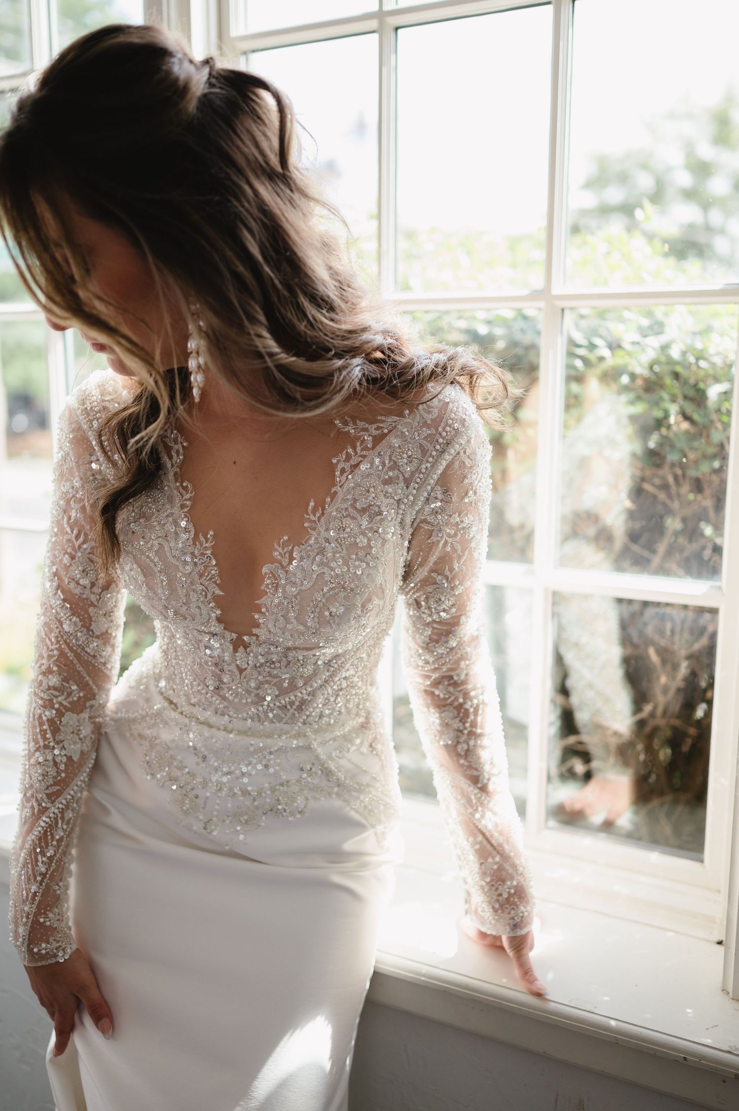Close-up details of an elegant white lace wedding dress with long sleeves against a sunlit window backdrop.