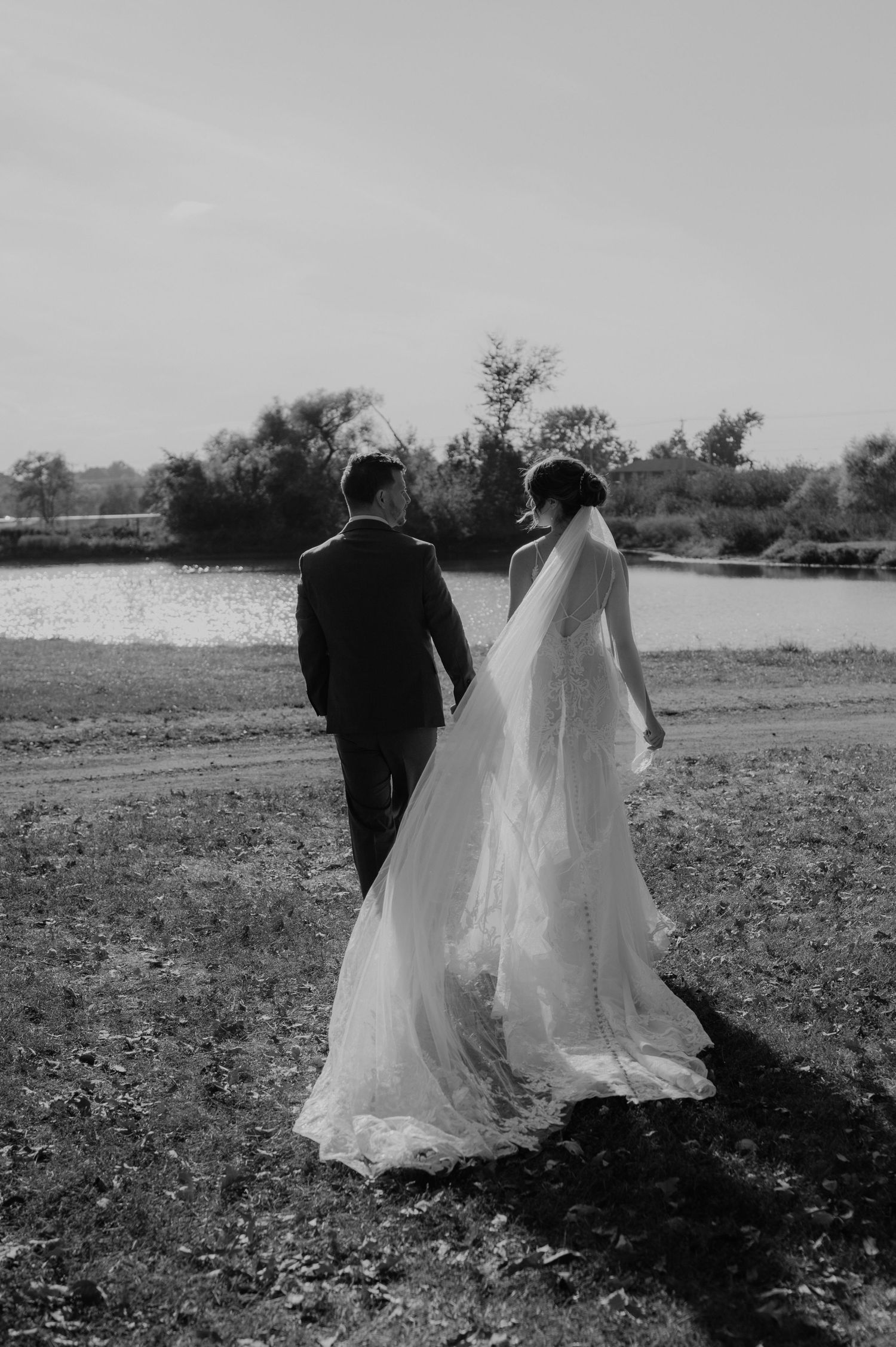 A black and white wedding photo showing couple walking by a lake with flowing veil trailing behind them in nature.