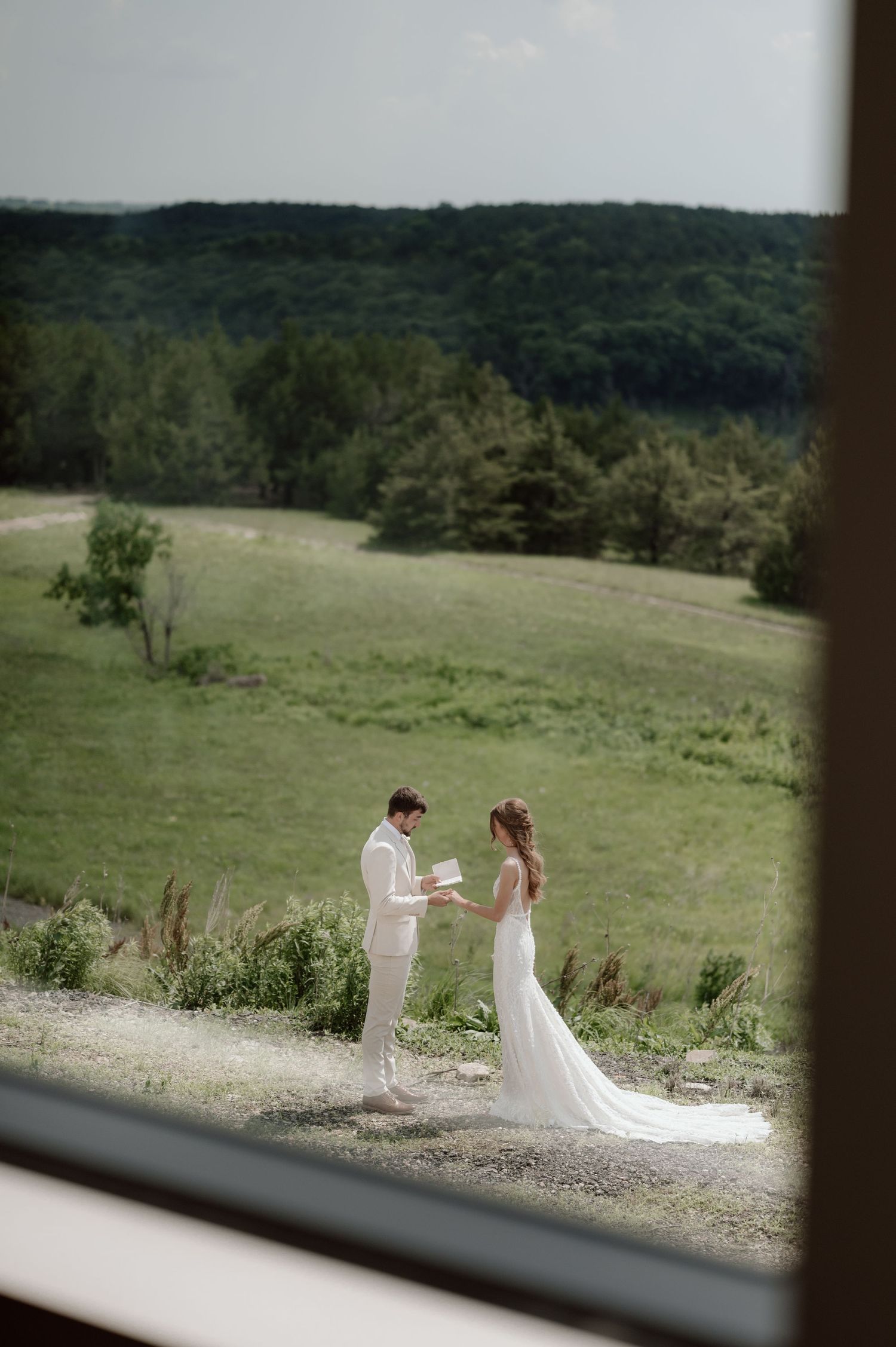 A couple in wedding attire stands together on a hilltop path with rolling green hills and forest in the background.