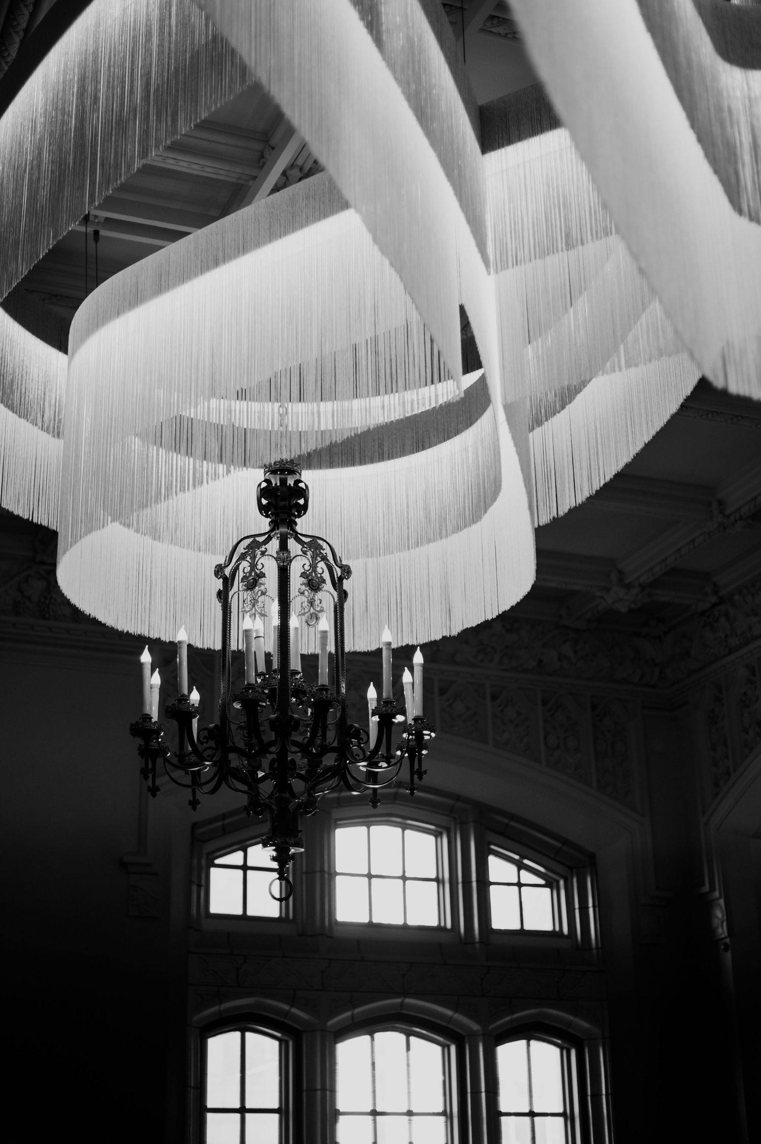 Elegant black chandelier hangs beneath flowing white fabric draping across a vaulted ceiling with arched windows.
