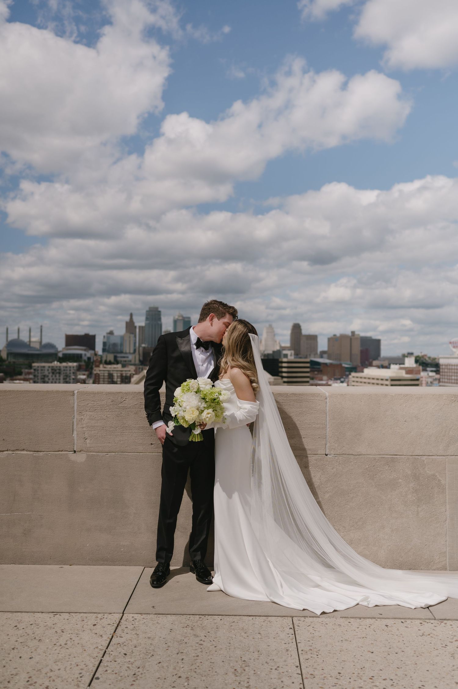 A romantic wedding couple shares intimate moments against a city skyline backdrop from a rooftop setting.