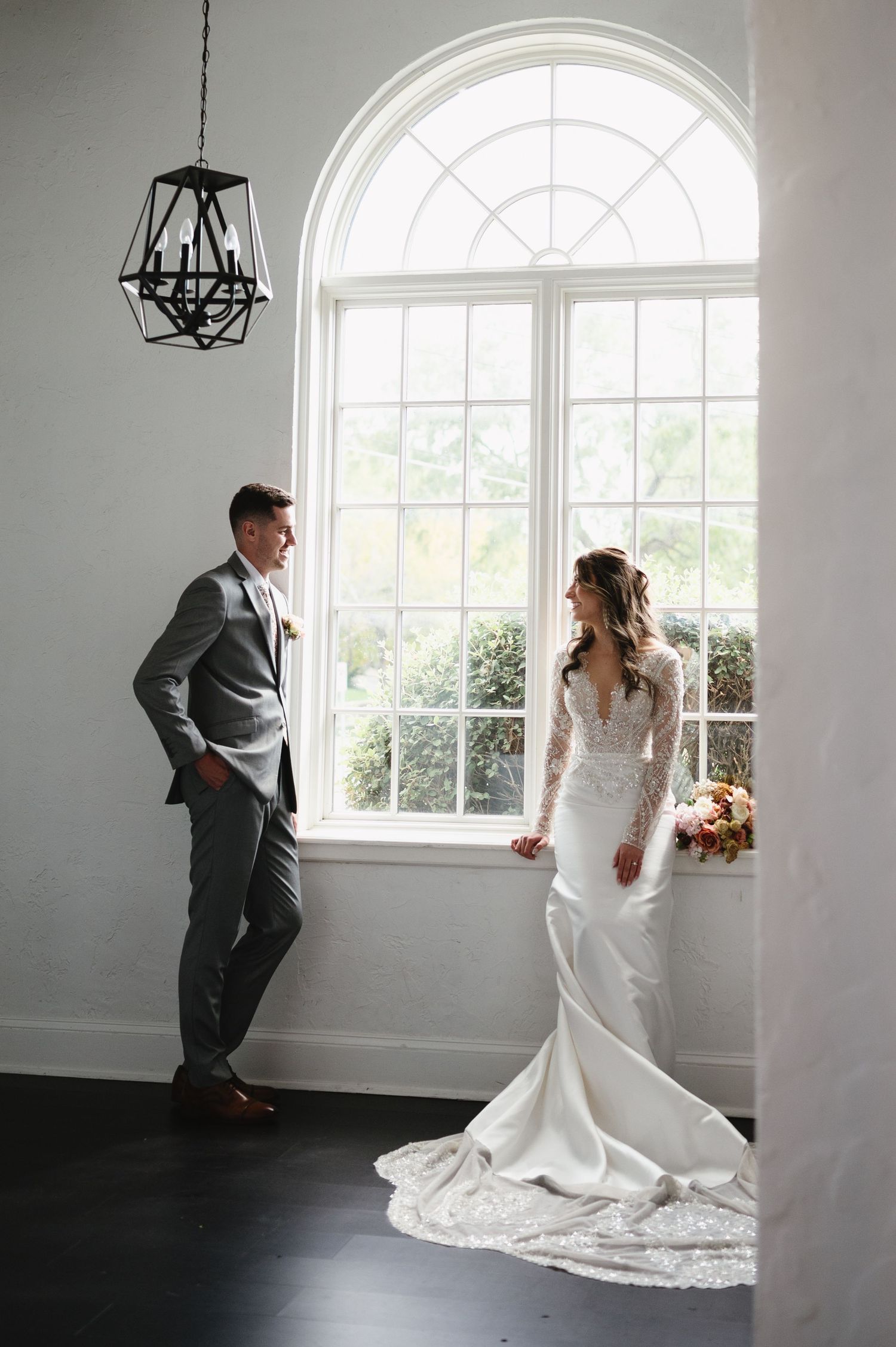 A couple in elegant wedding attire stands by a large arched window with natural light streaming in, creating a romantic scene.