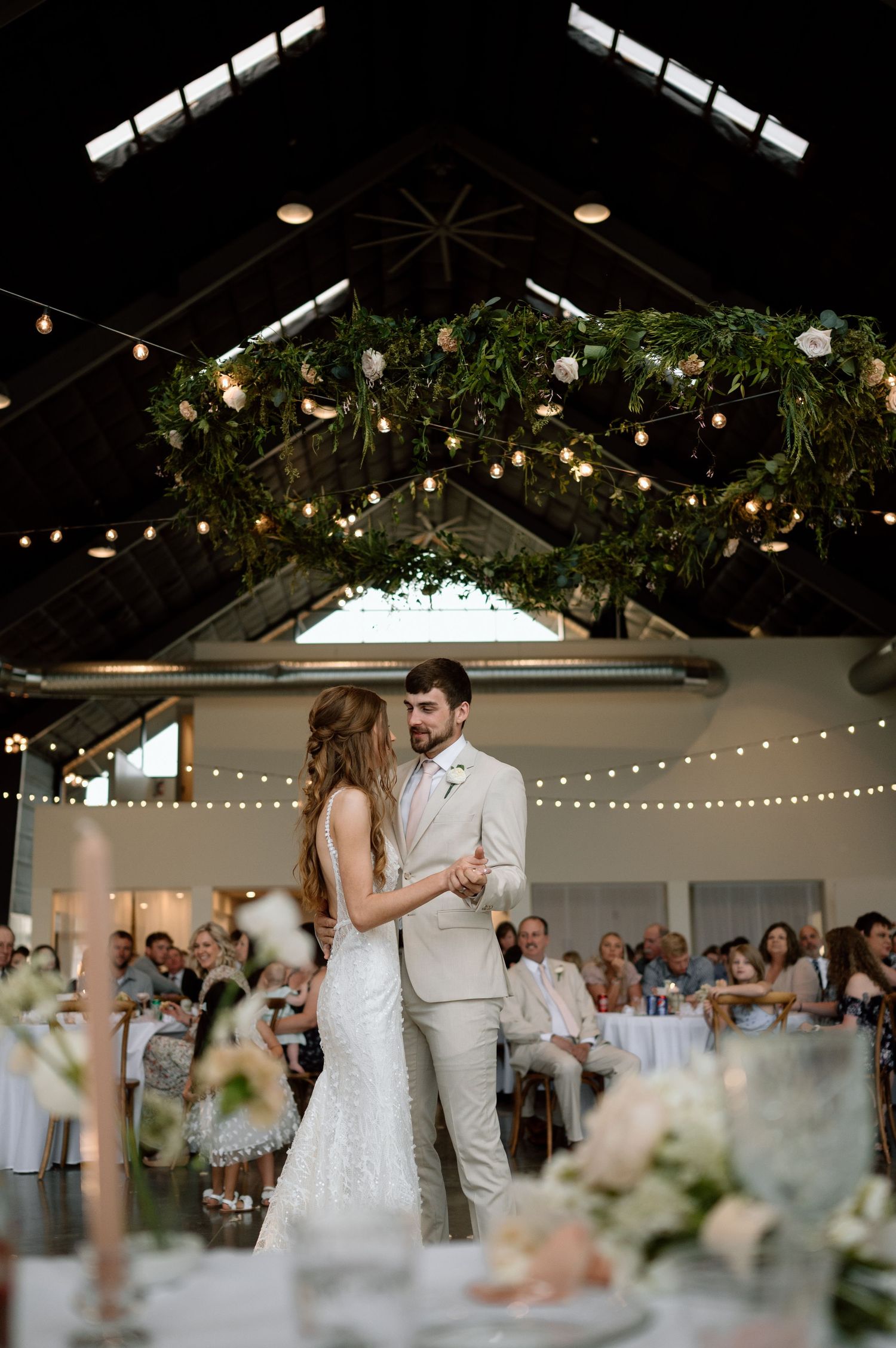 A romantic wedding reception in a barn-style venue with string lights, greenery, and floral decorations overhead.