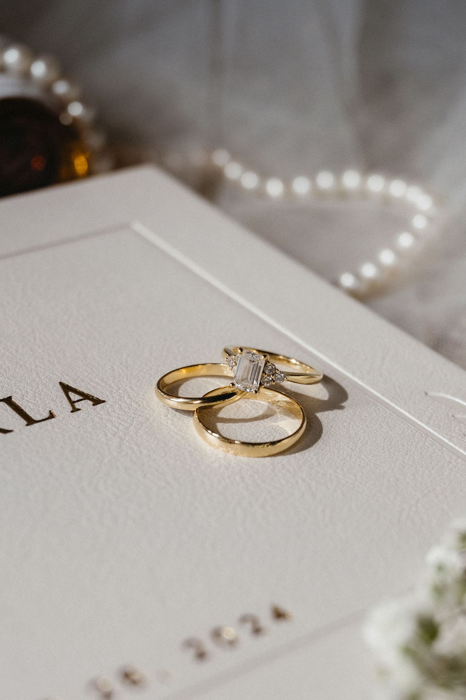 Gold wedding rings and engagement ring with diamond resting on elegant white paper with string of pearls.