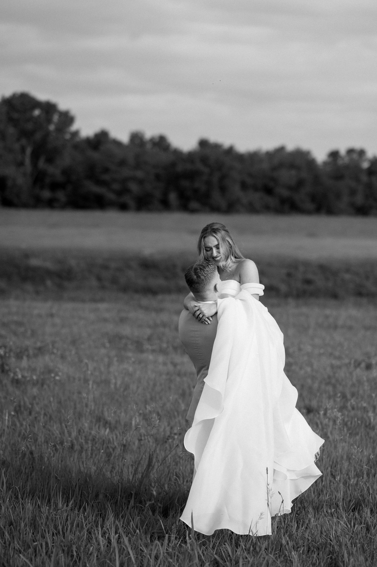A bride in a flowing white gown stands in a serene field with trees in the background at dusk.
