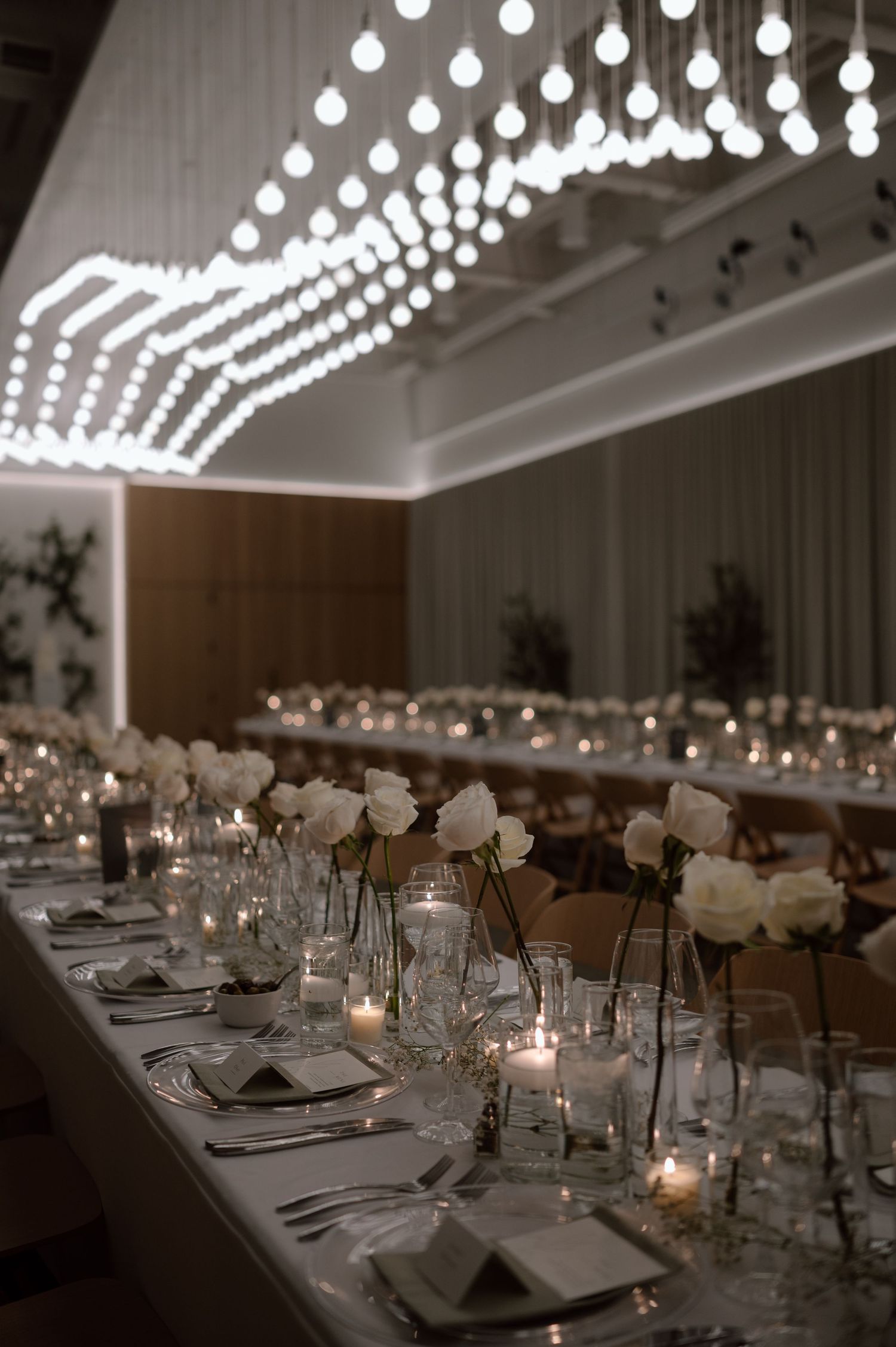 String lights create an elegant ambiance above long dining tables set with white linens and glowing candles in a reception hall.