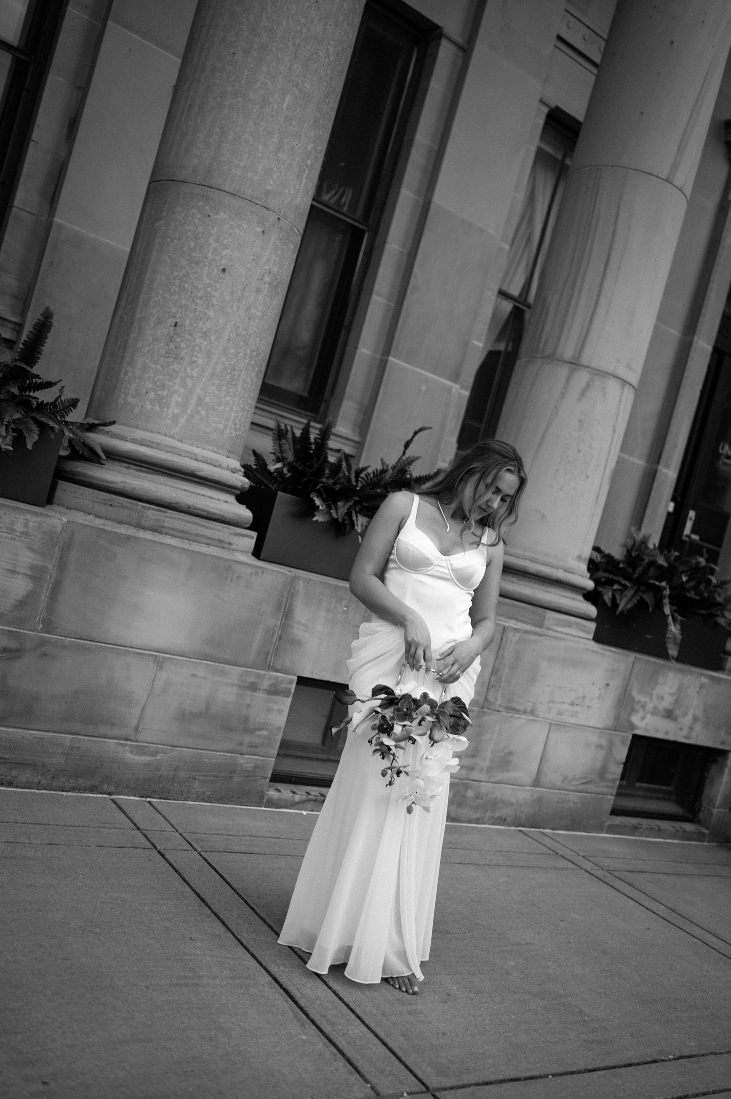 A bride in a white wedding dress poses on a concrete walkway flanked by classical stone columns.