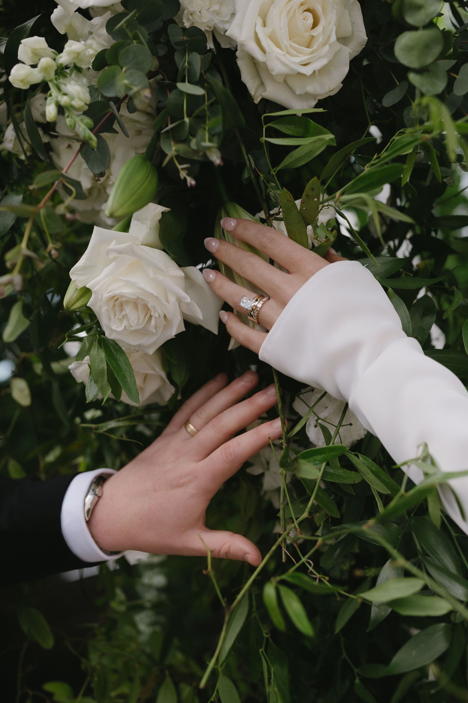 A hand with rings rests against a lush floral arrangement of white roses and greenery on a backdrop.