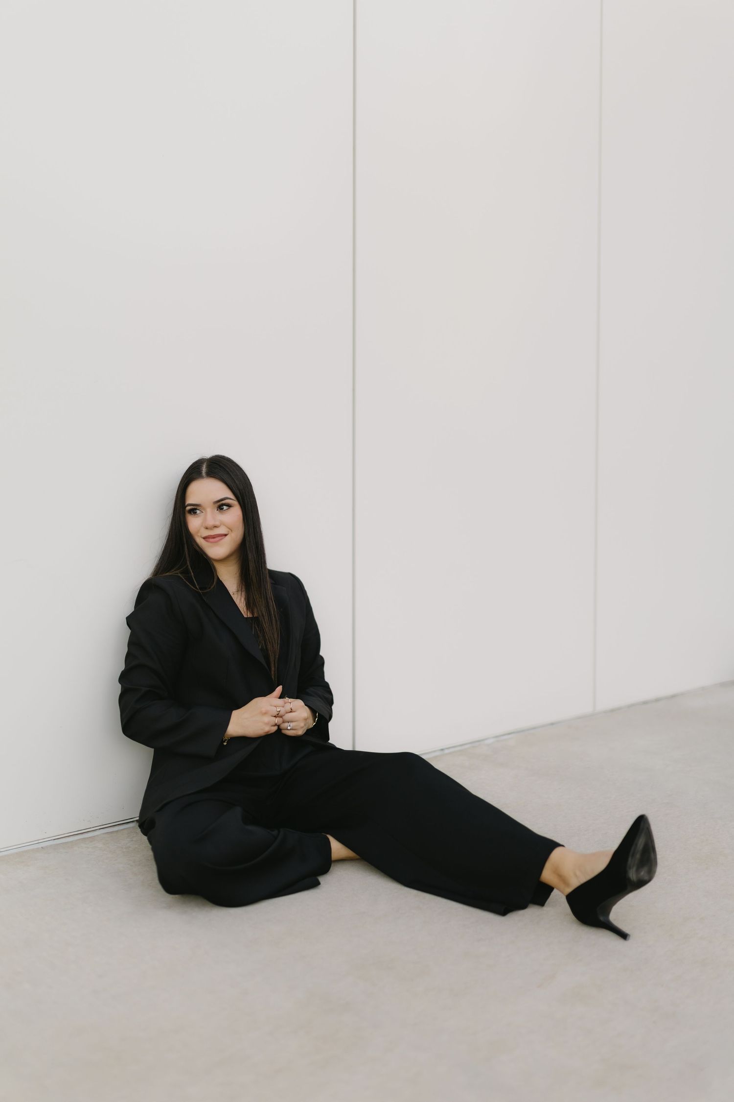 Person in all black attire sits on floor against white wall in minimalist setting with heels and long dark coat.
