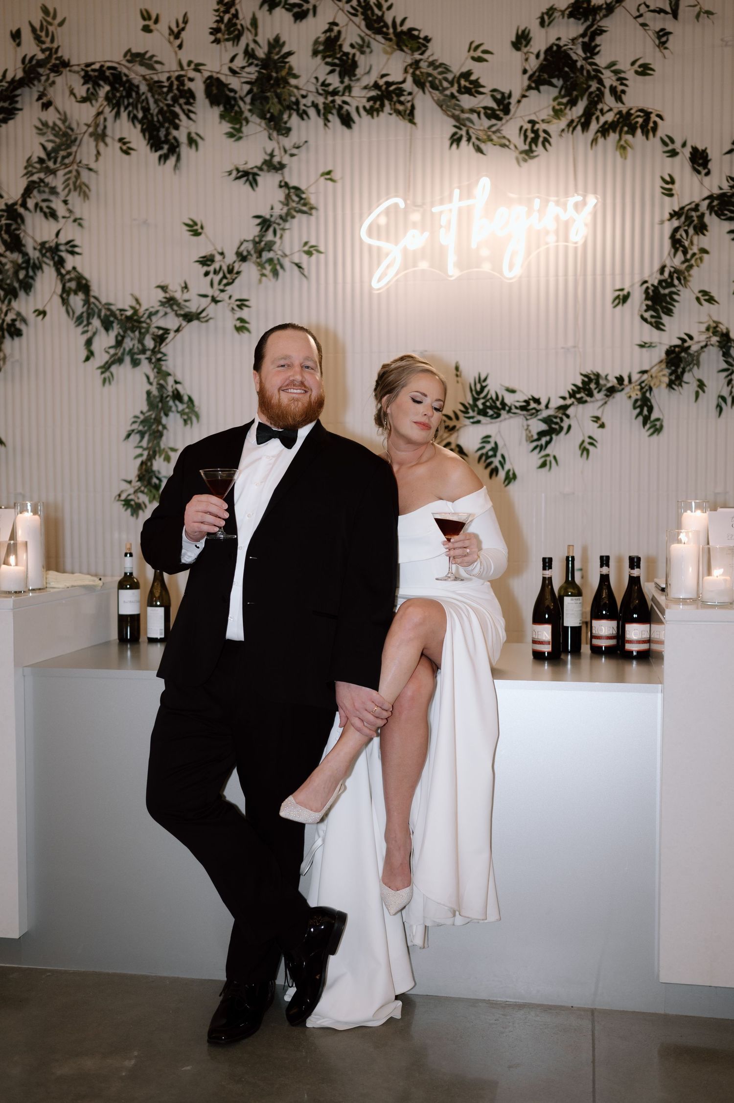 A couple in formal attire pose and laugh together in an elegant white reception space with hanging greenery.