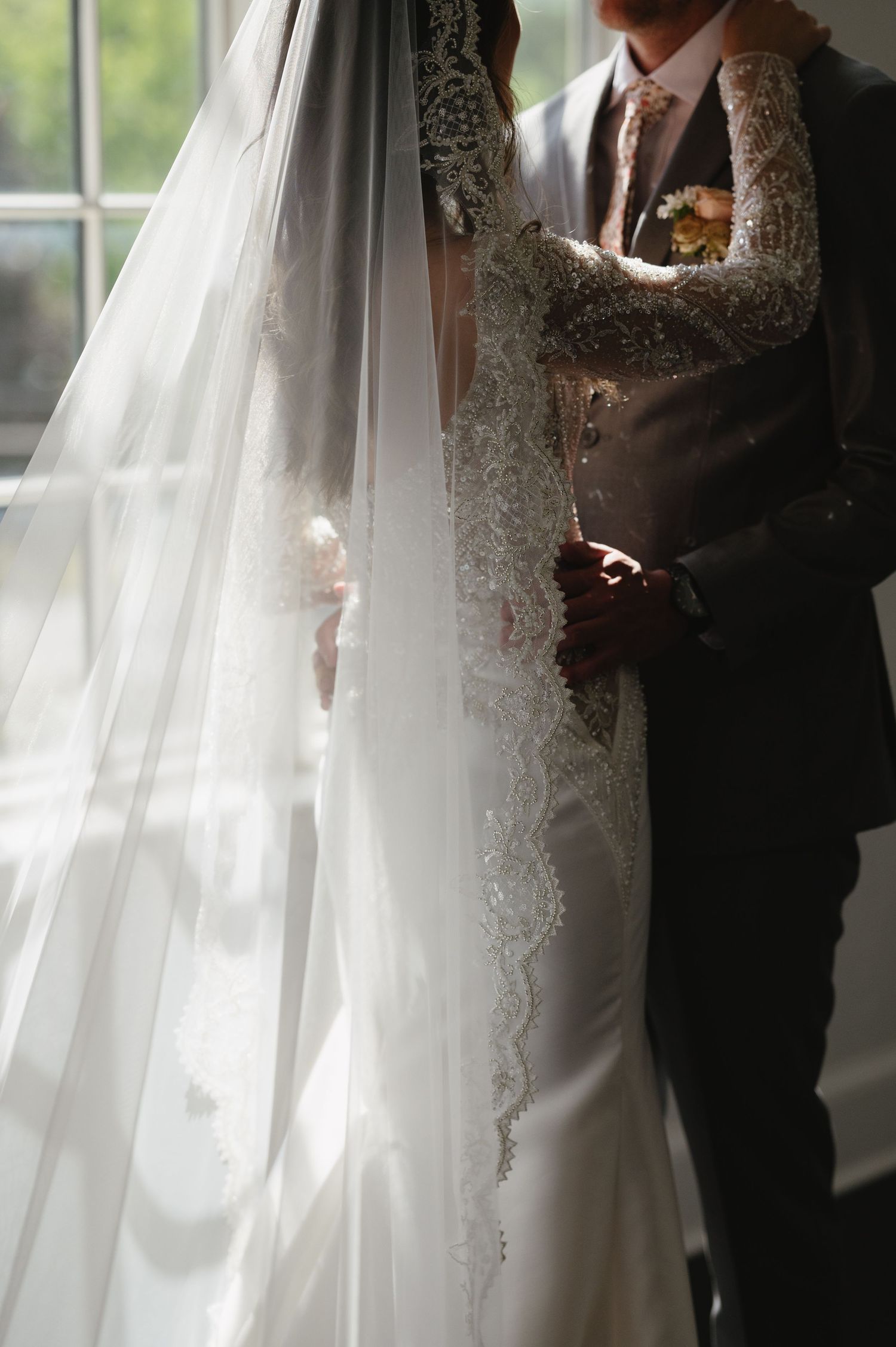 A wedding couple in an elegant white lace dress and gray suit stand by a window with a flowing veil in soft natural light.