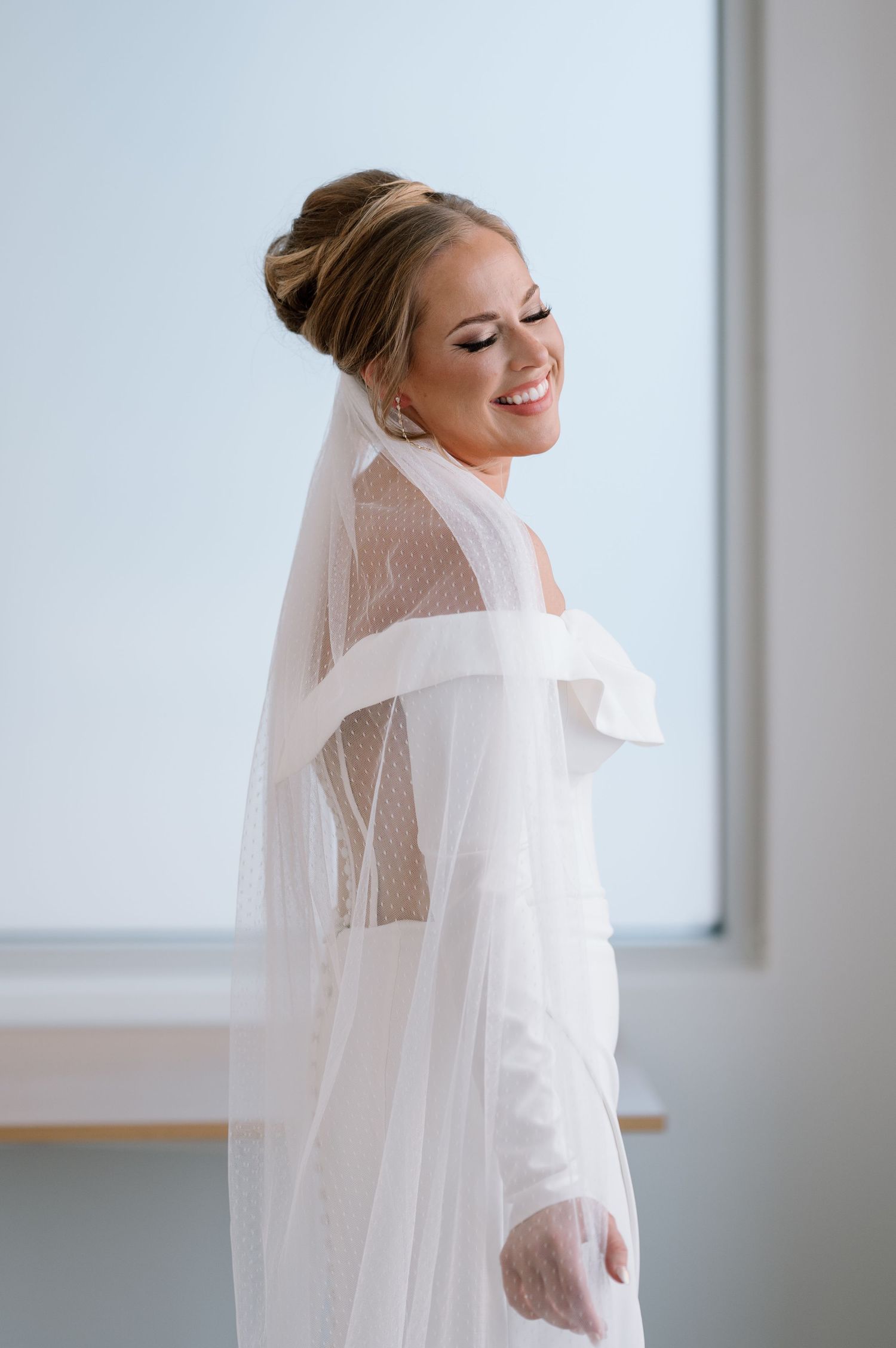 A bride in a sheer robe and veil smiles joyfully while standing near a window in natural light.