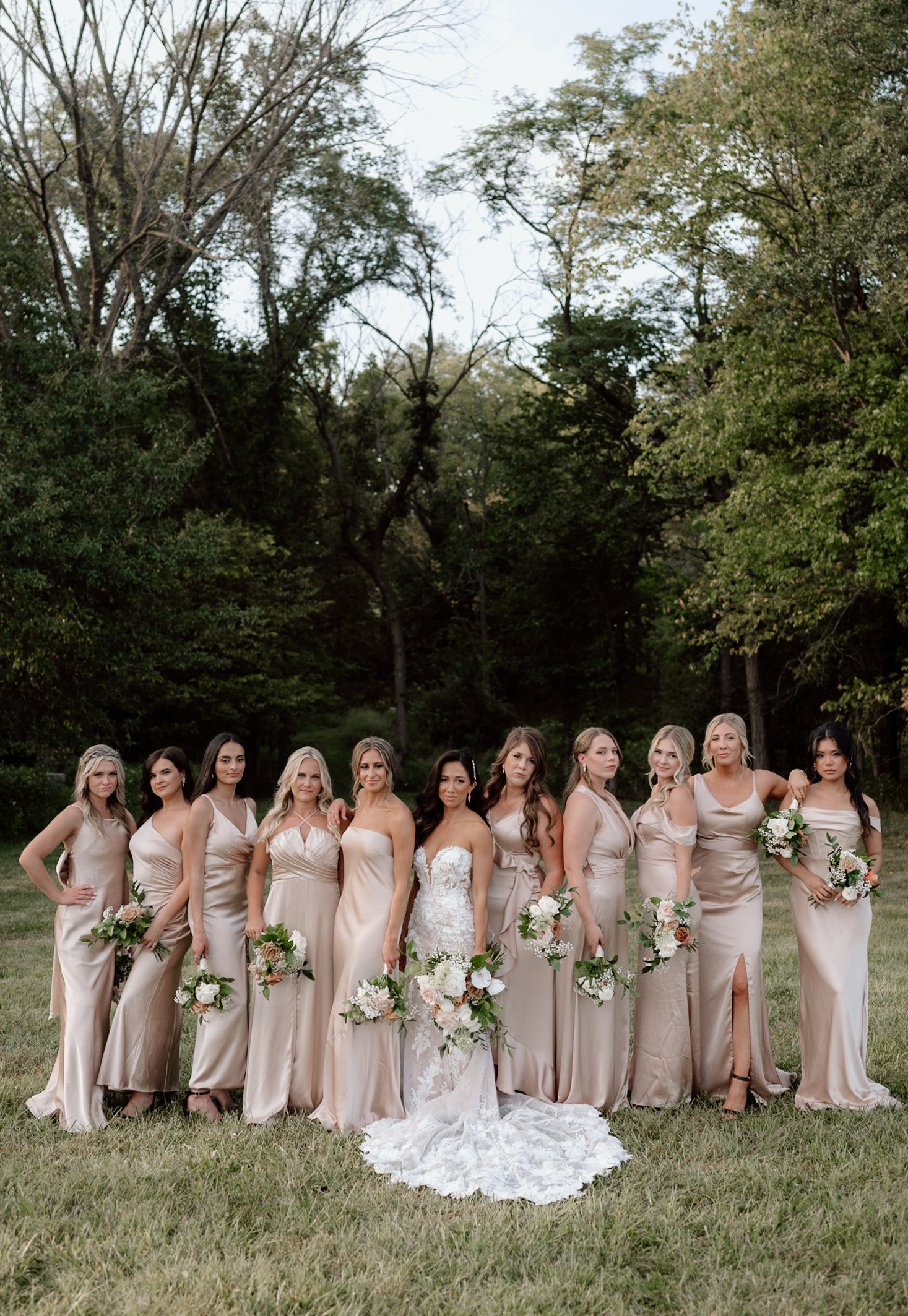 A bridal party wearing blush pink bridesmaid dresses poses together on grass with trees in the background.