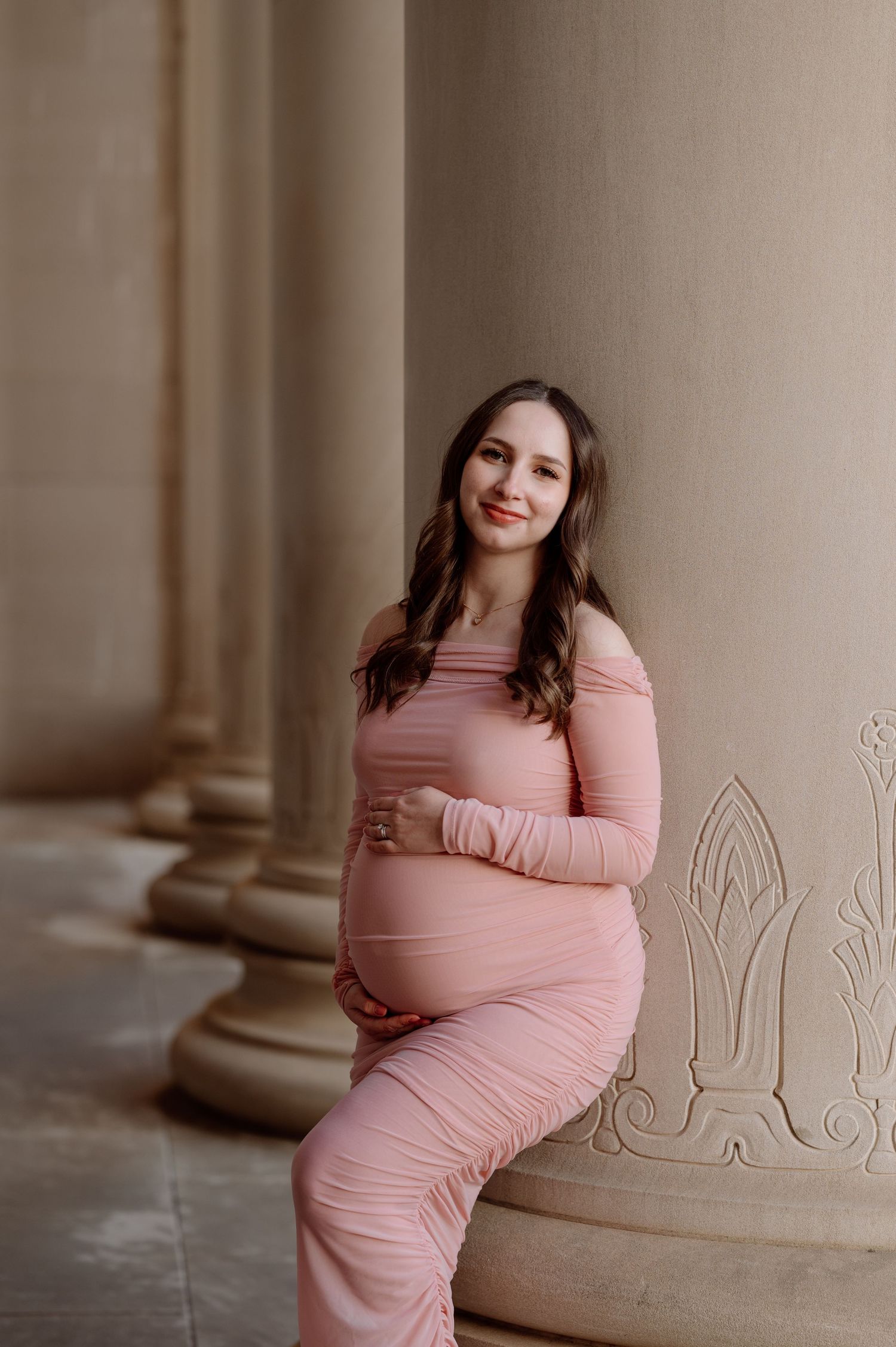 An expectant mother in an off-shoulder pink dress poses elegantly against classic stone columns in a soft, romantic setting.