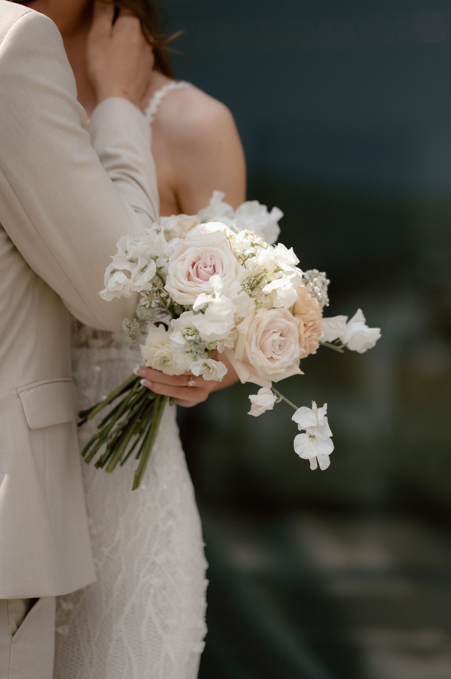 A delicate blush and white wedding bouquet held against a cream suit and lace dress during an intimate moment.