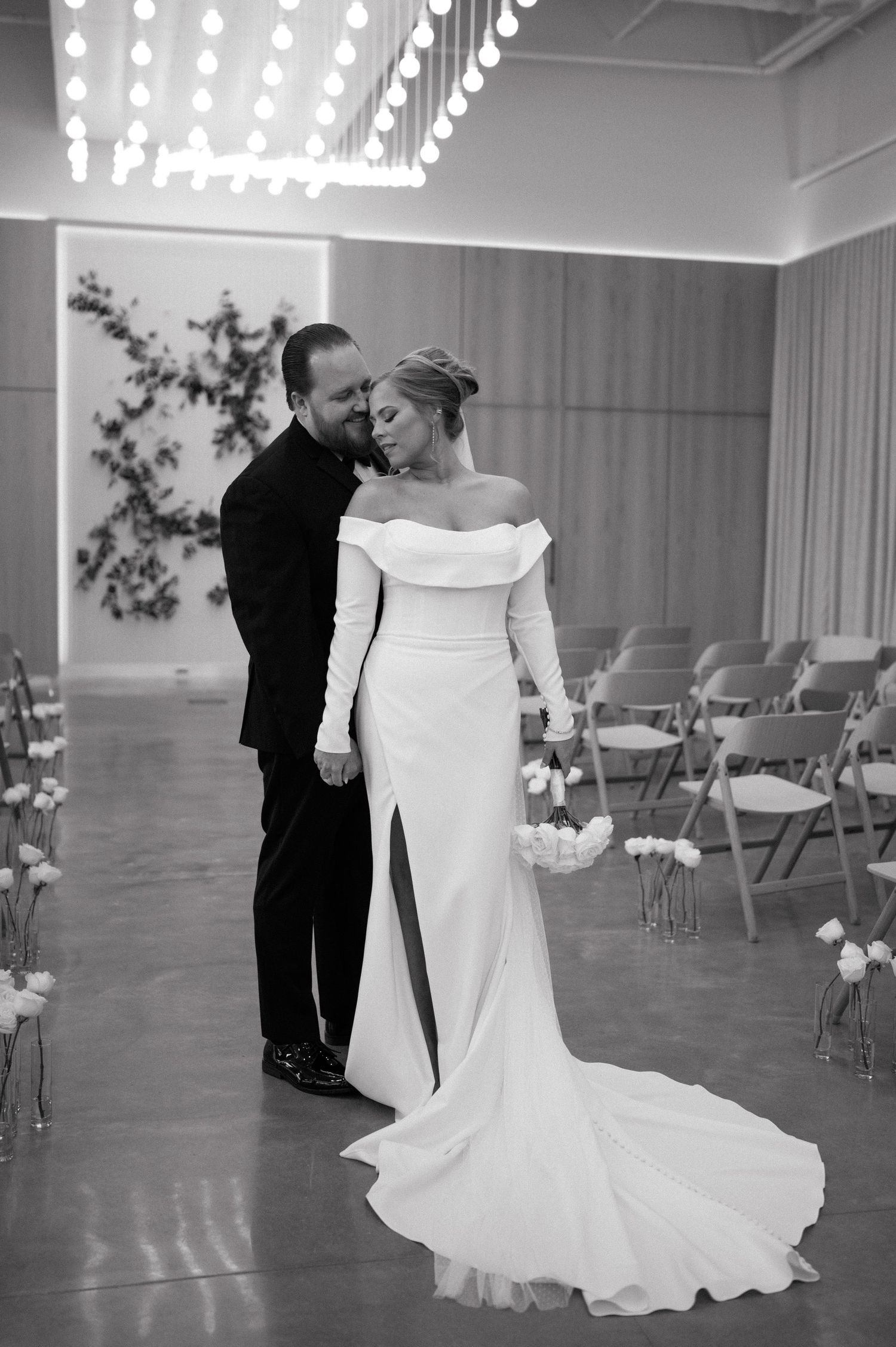 Black and white photo of a couple sharing an intimate moment at indoor wedding ceremony with chairs and florals.