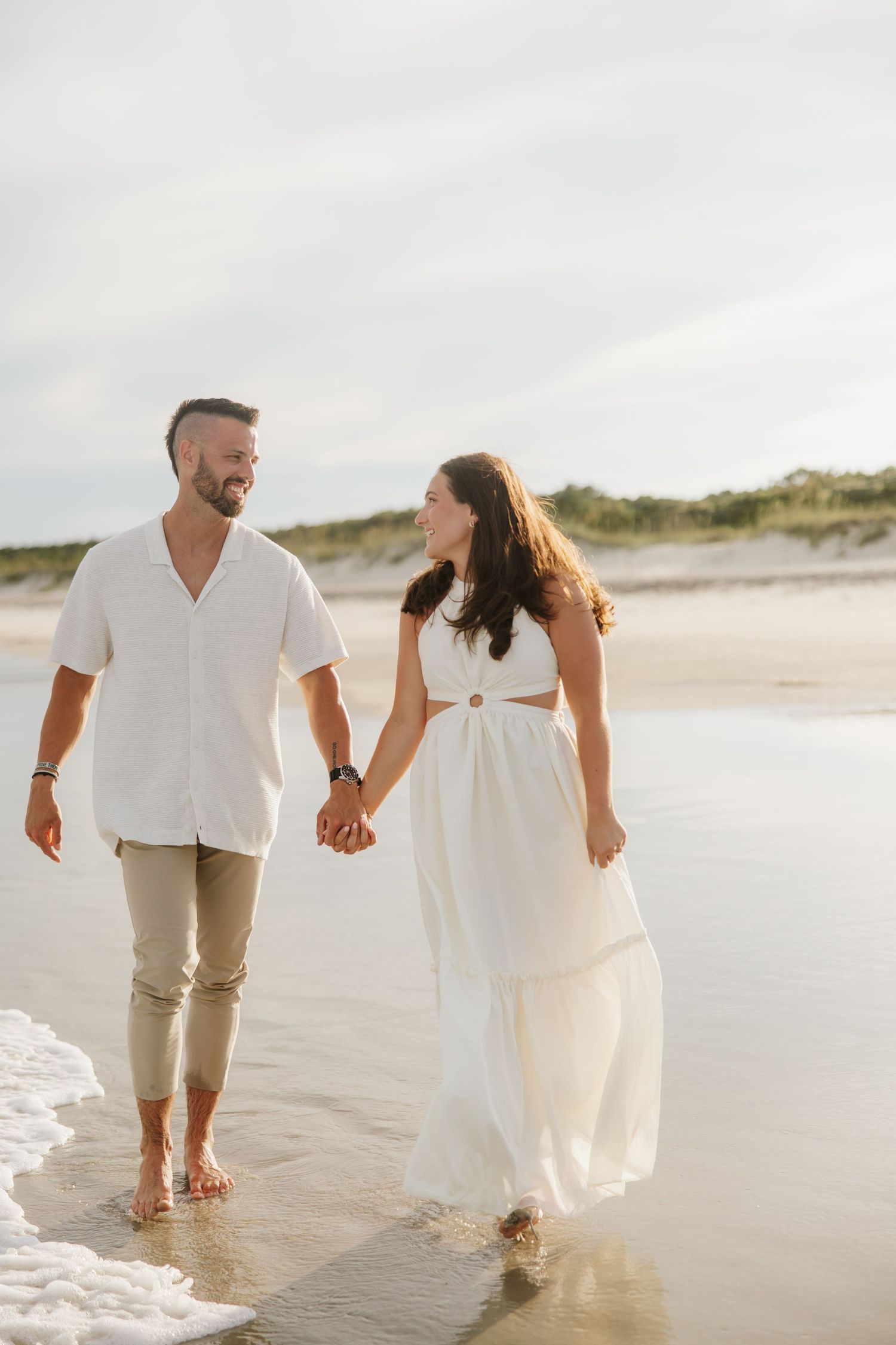 Happy couple walking hand in hand along a sunny beach, dressed in white outfits, smiling at each other.