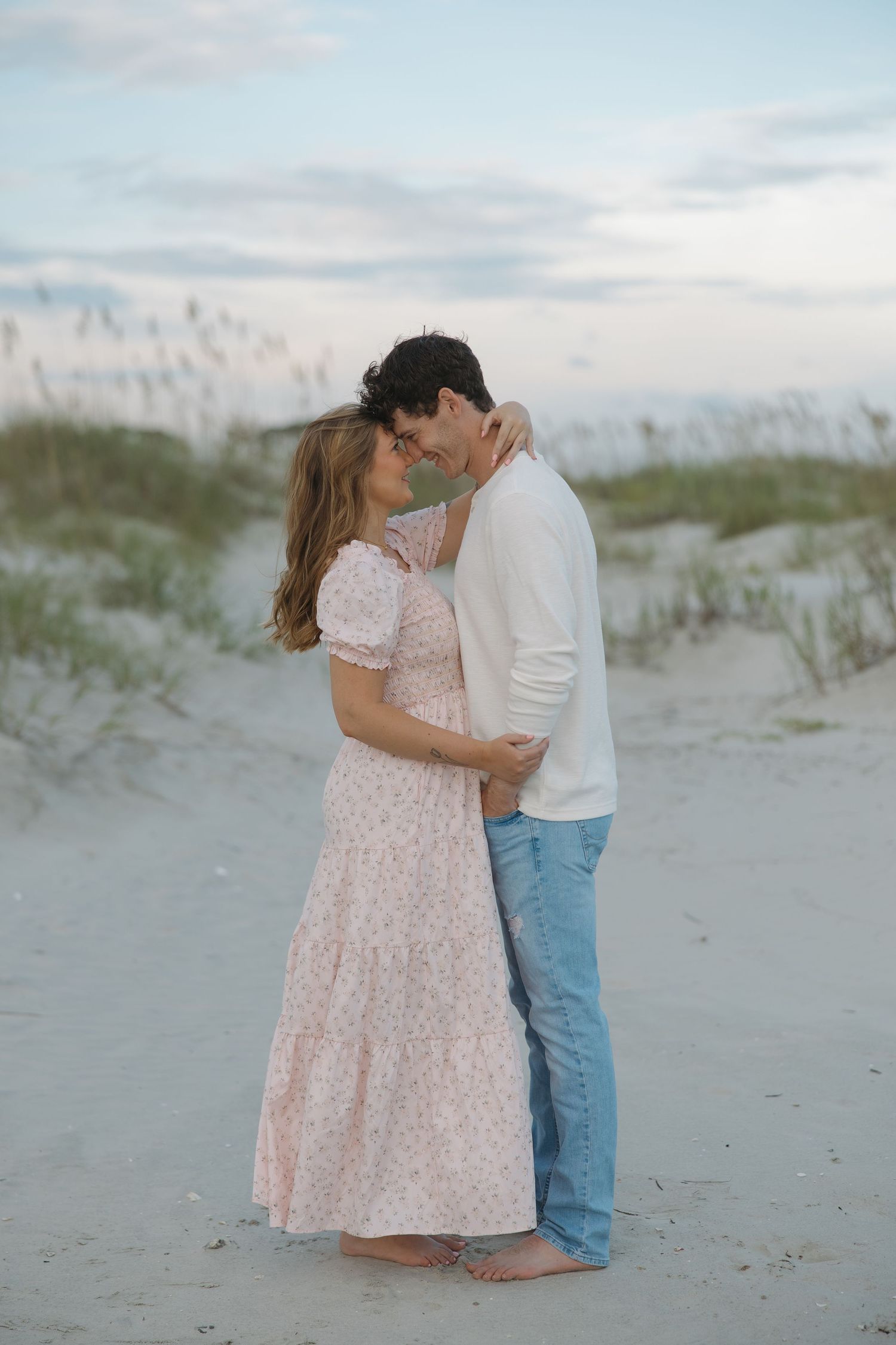 A couple embraces romantically on a sandy beach at dusk, woman in floral dress, man in white shirt and jeans.
