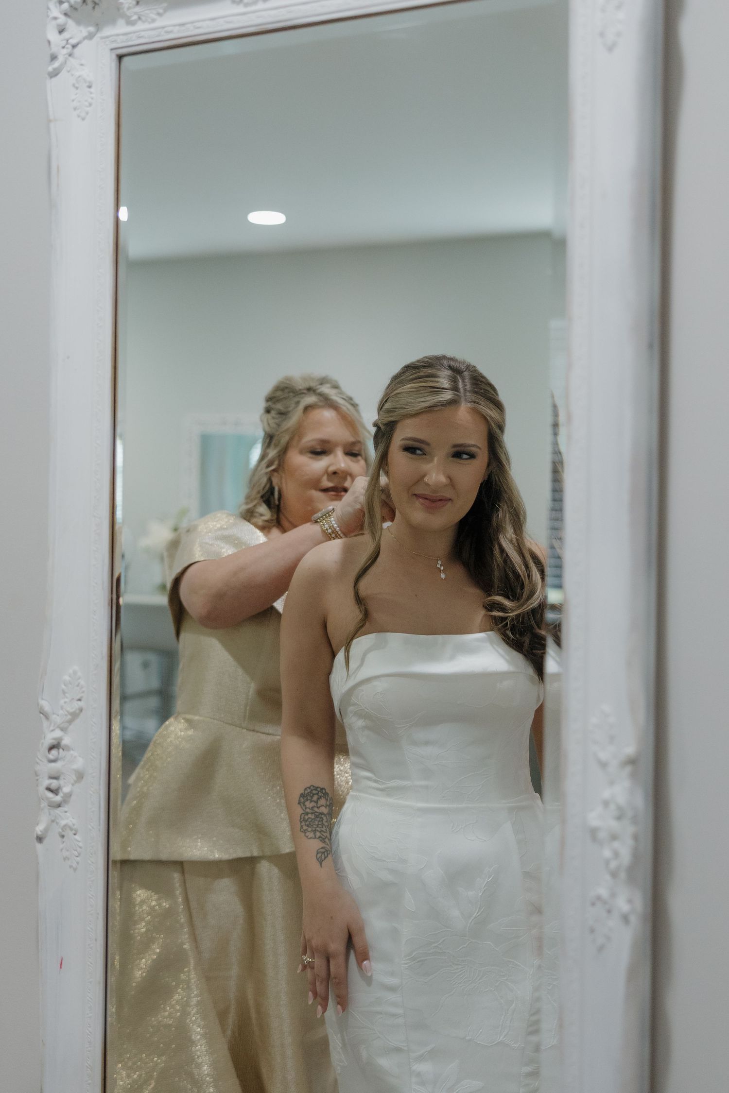 A bride in a white strapless gown is reflected in an ornate white mirror as a woman in a gold dress helps her get ready.