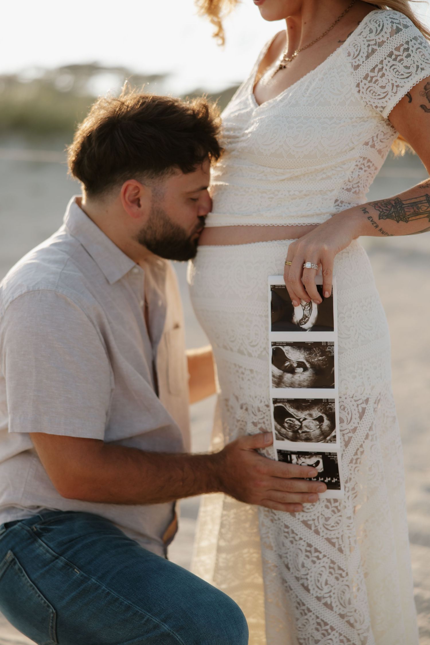 Man kisses pregnant woman's belly while holding ultrasound photos on a sunny beach during a maternity photoshoot.