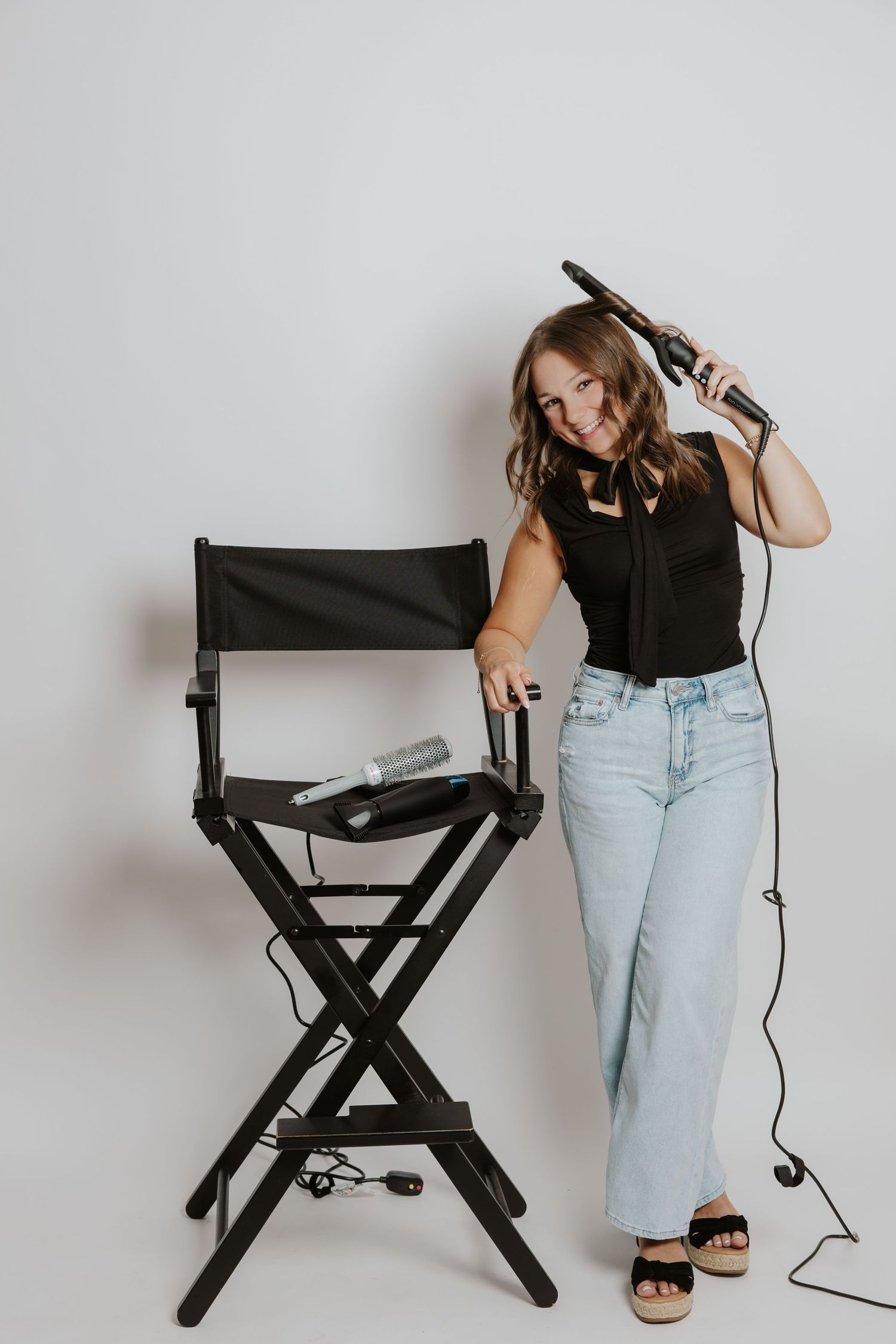 Female hairstylist using curling iron beside black director chair with hair tools on white background.