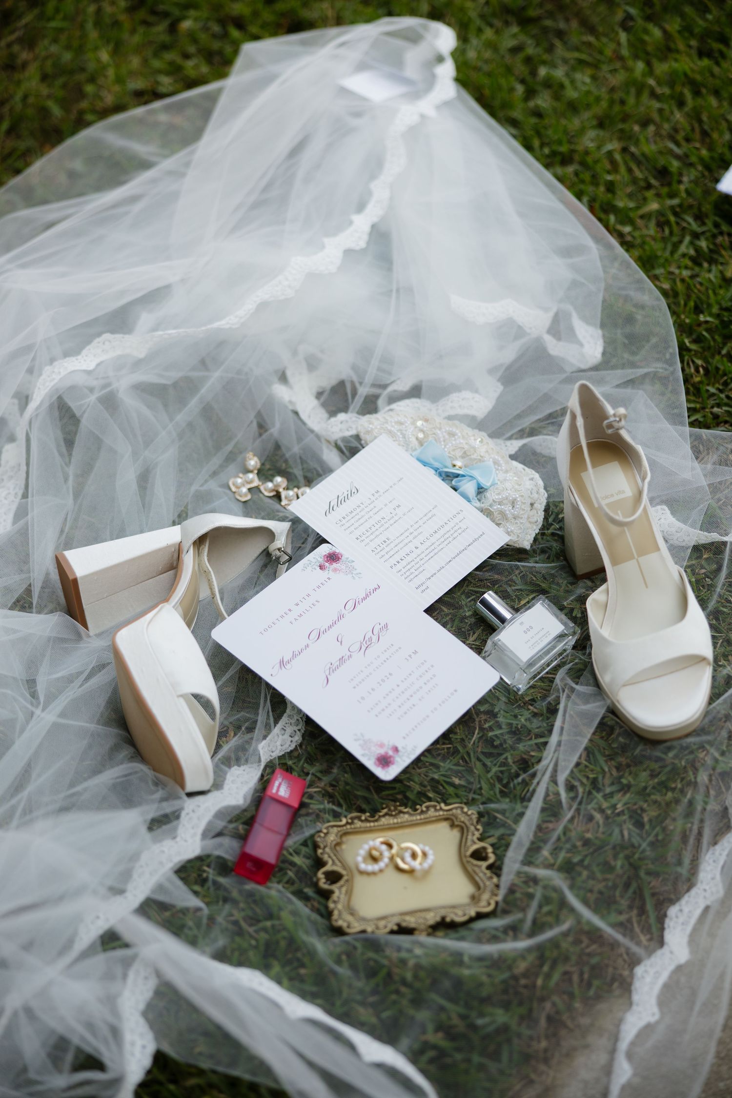 Bridal flat lay with a lace-edged veil, white heels, wedding invitation, perfume, red lipstick and rings on a gold tray.