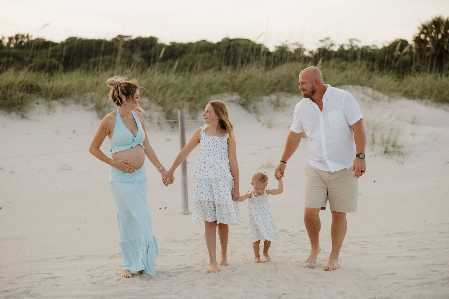 Pregnant woman in blue maxi dress holds hands with family on sandy beach with dunes and grass in background.