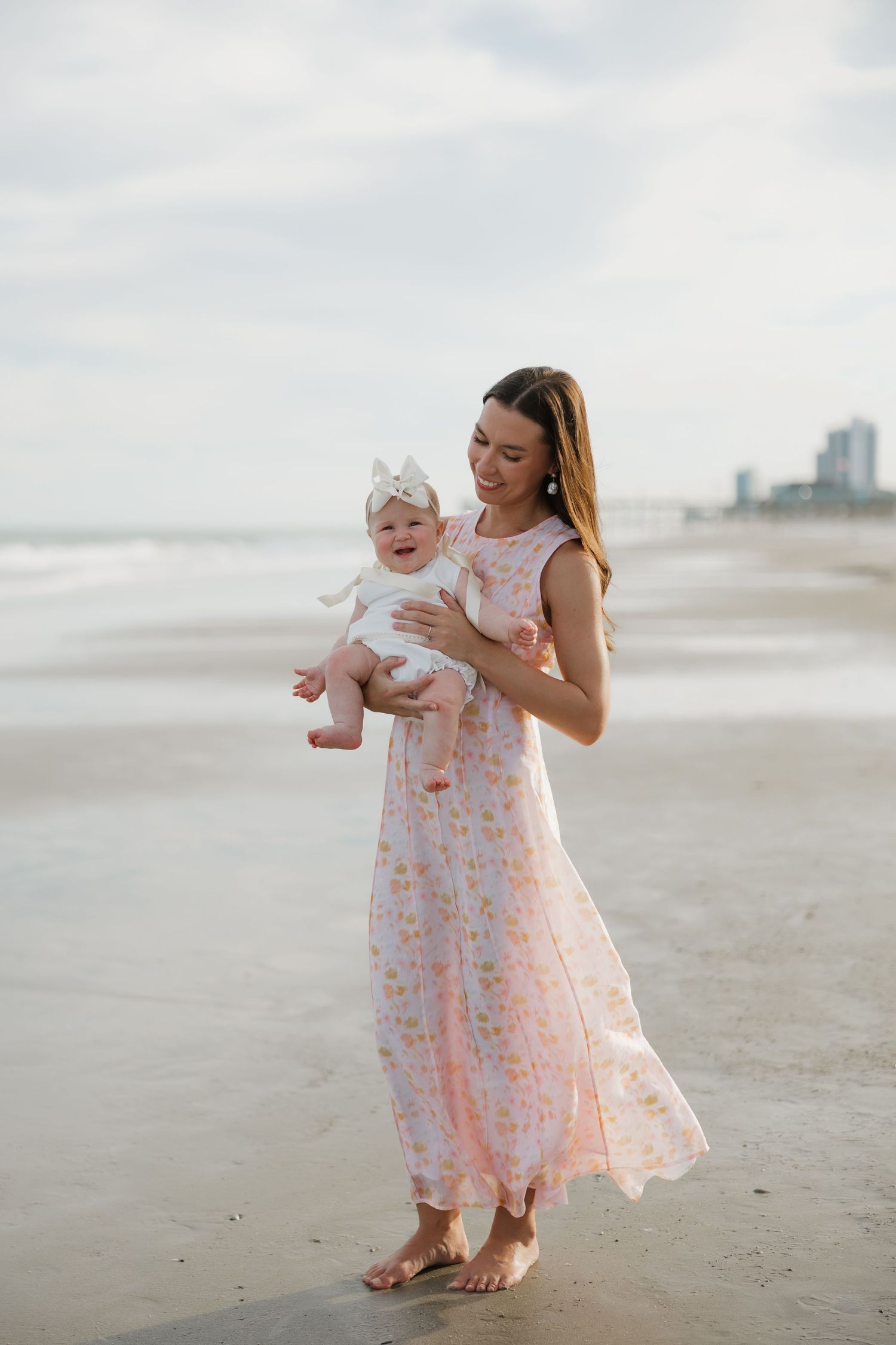 A mother in a floral pink maxi dress holds her baby girl on a sandy beach.