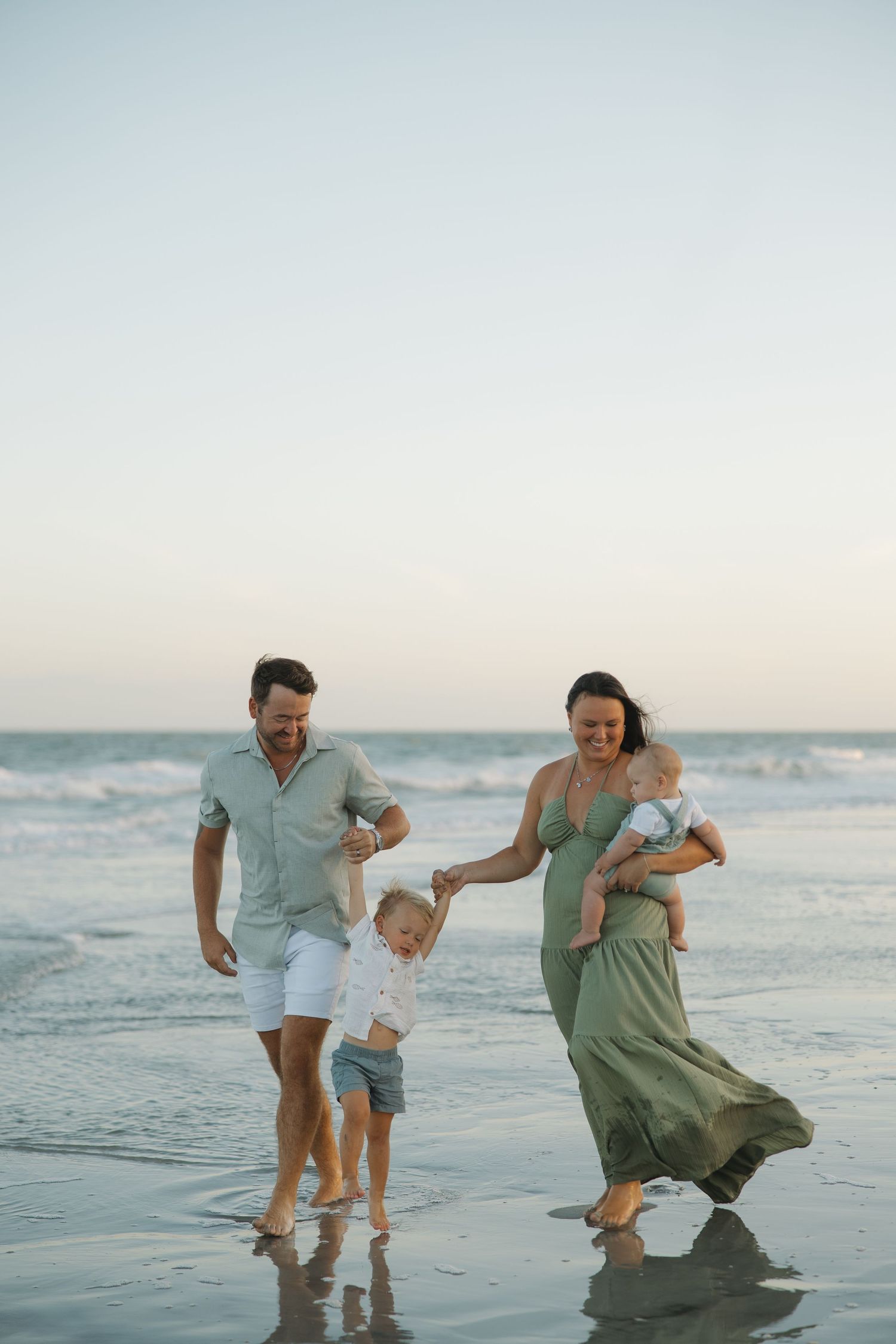 Happy family of four walking on a beach at sunset, parents with toddler and baby enjoying the ocean shore.