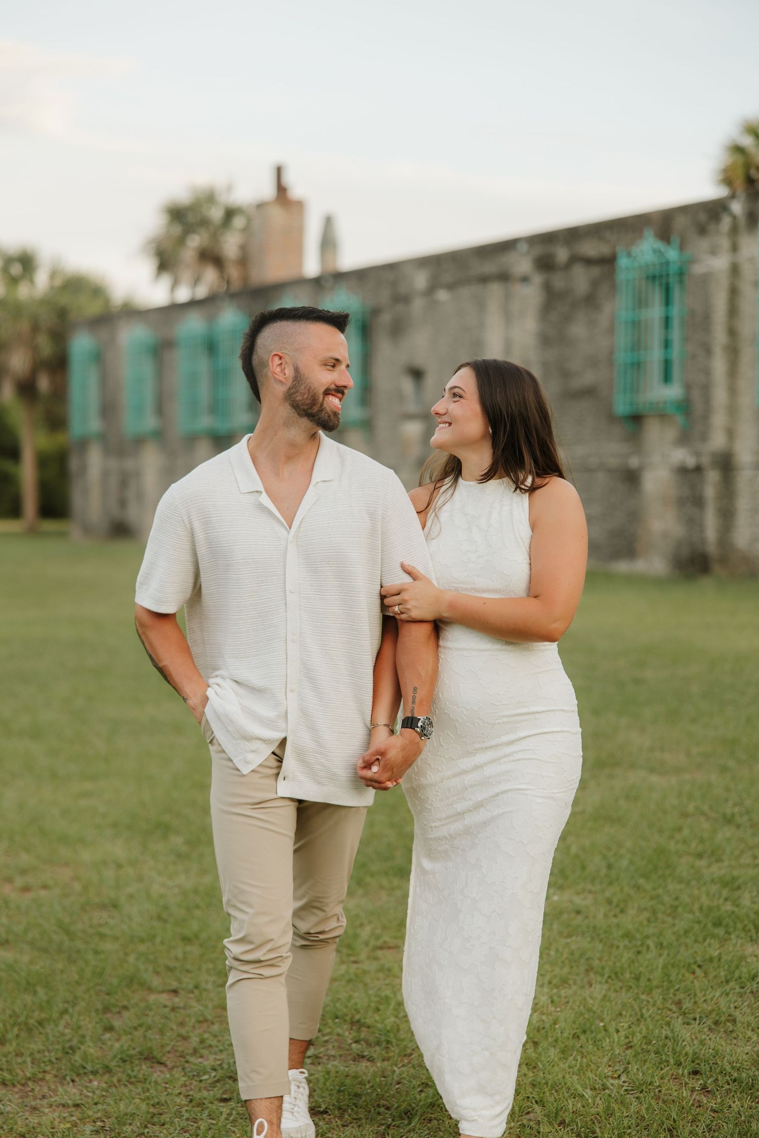 A smiling couple in white outfits walks hand in hand on green grass near a historic stone building.
