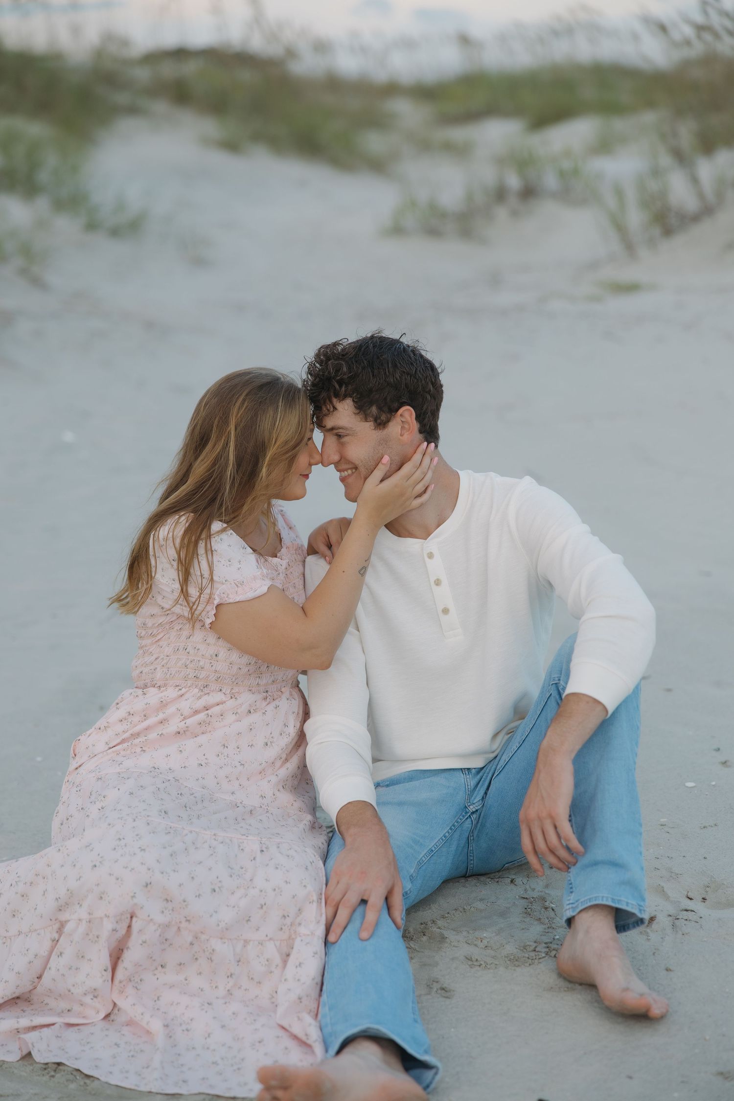 Couple sitting on sandy beach dunes, woman in floral dress touching mans face as they lean in close.