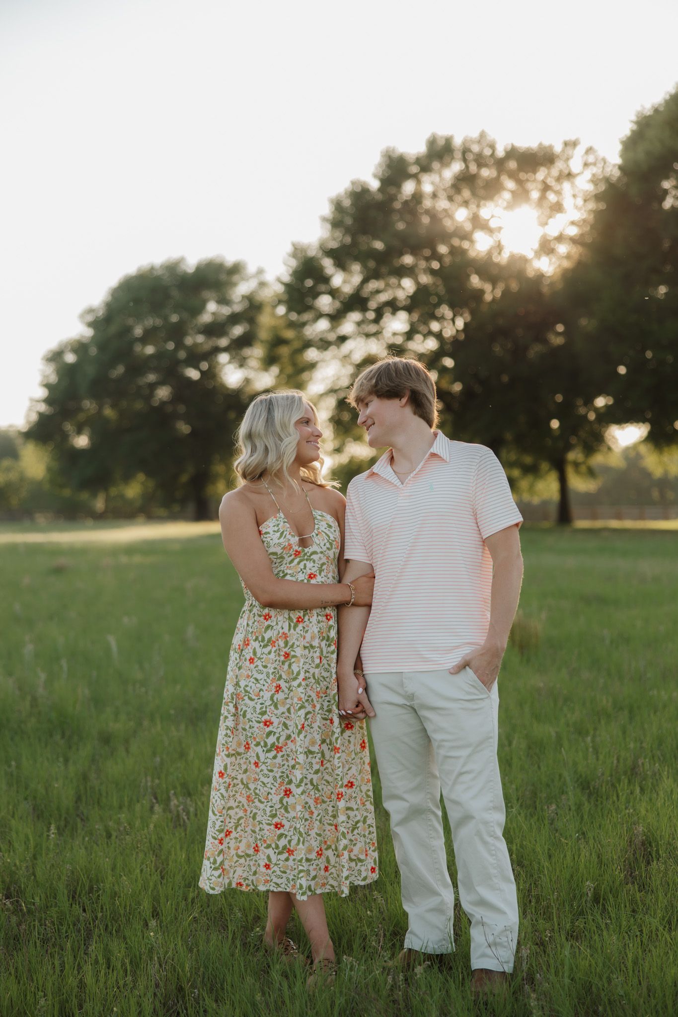 Couple holding hands in a sunlit green field, woman in floral dress and man in pink polo smiling at each other.