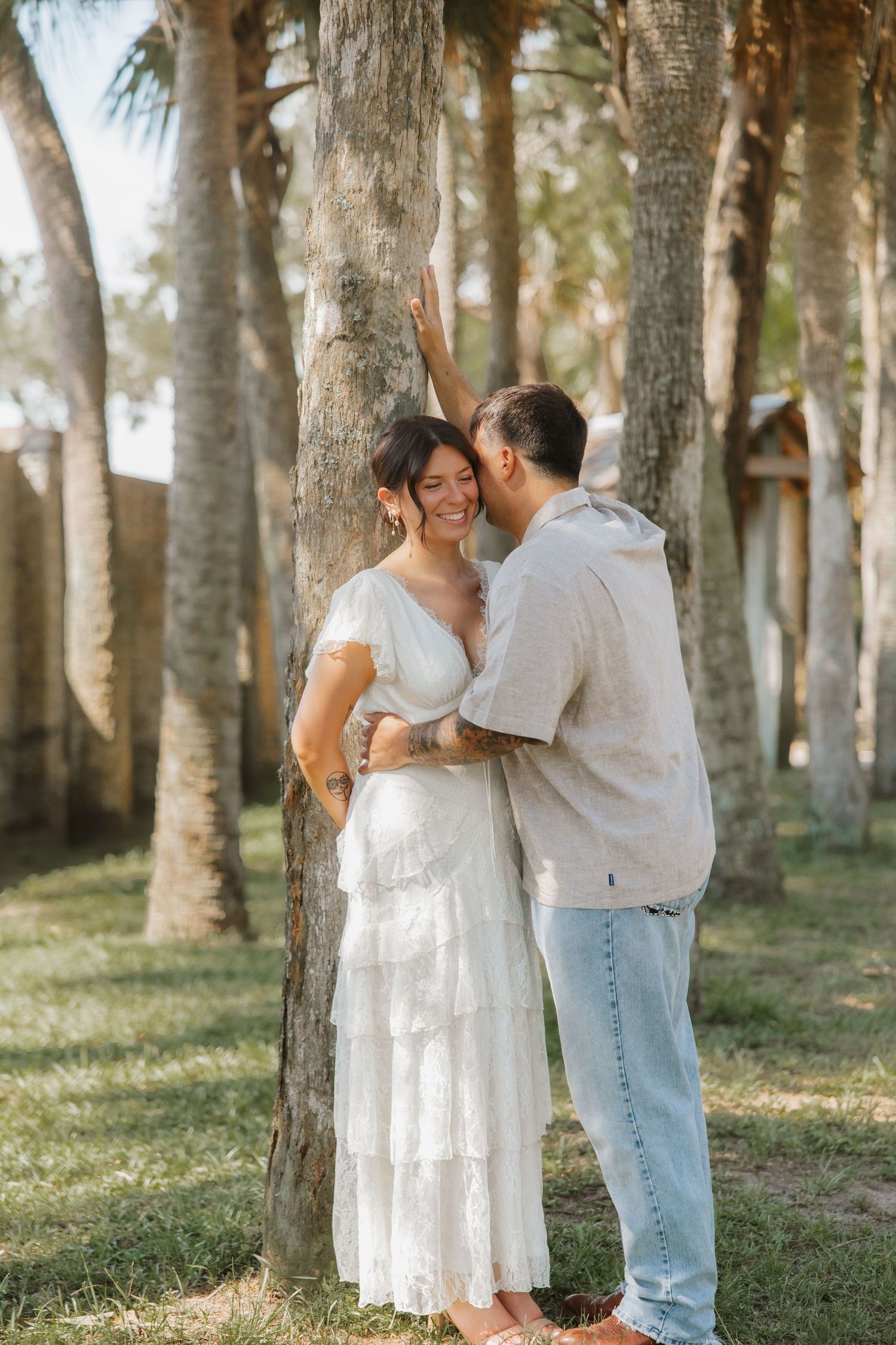 Couple embracing near palm trees, woman in white tiered dress smiling as man whispers in her ear outdoors.