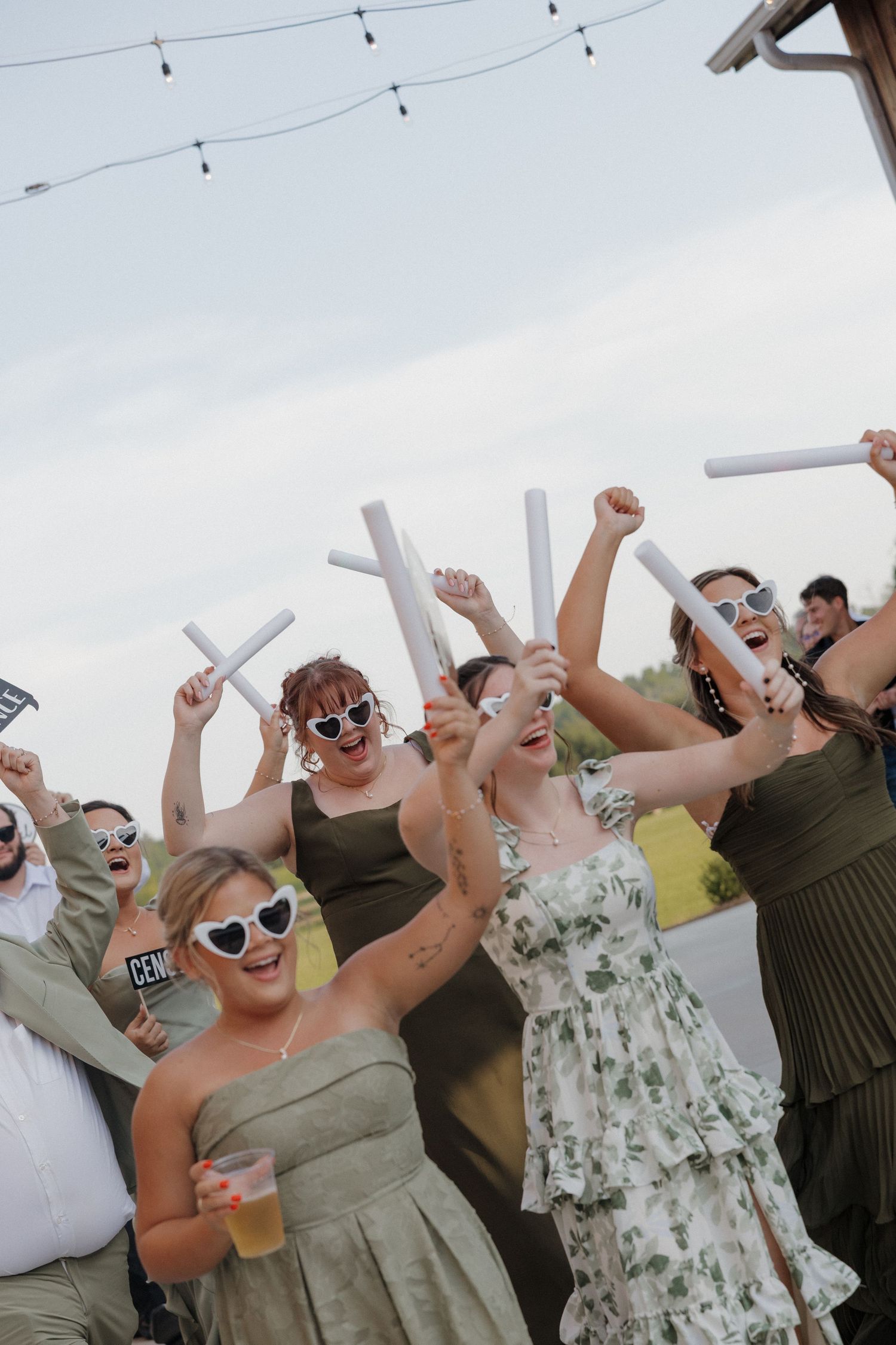 Smiling women in sage green dresses and white sunglasses cheer outdoors at a celebration, raising foam sticks in the air.