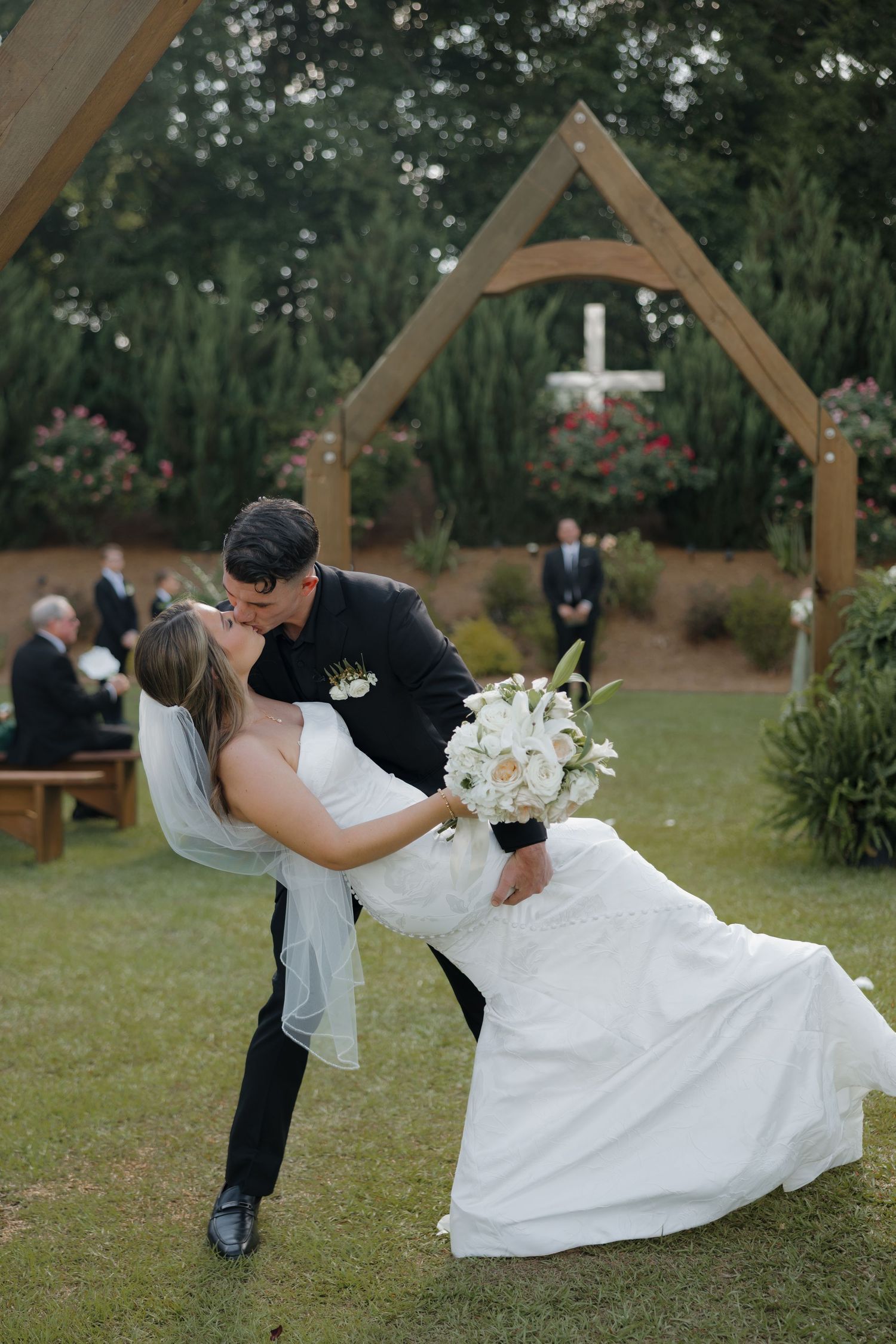 Groom in a black suit dips and kisses his bride in a white gown holding a bouquet near a wooden outdoor ceremony arch with a cross.
