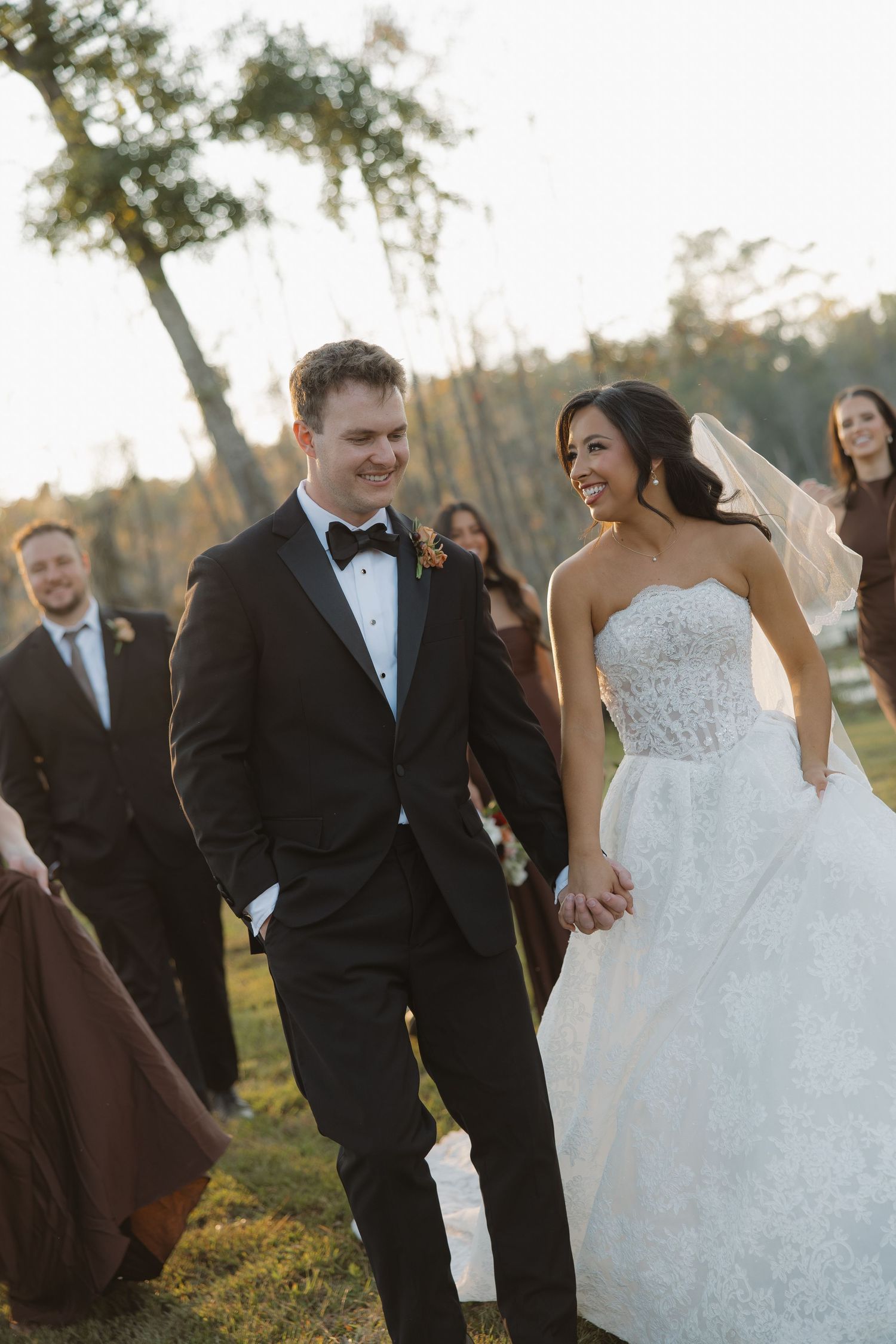 Smiling bride in a strapless lace wedding gown holds hands with a groom in a black tuxedo and bow tie as their wedding party walks outdoors.