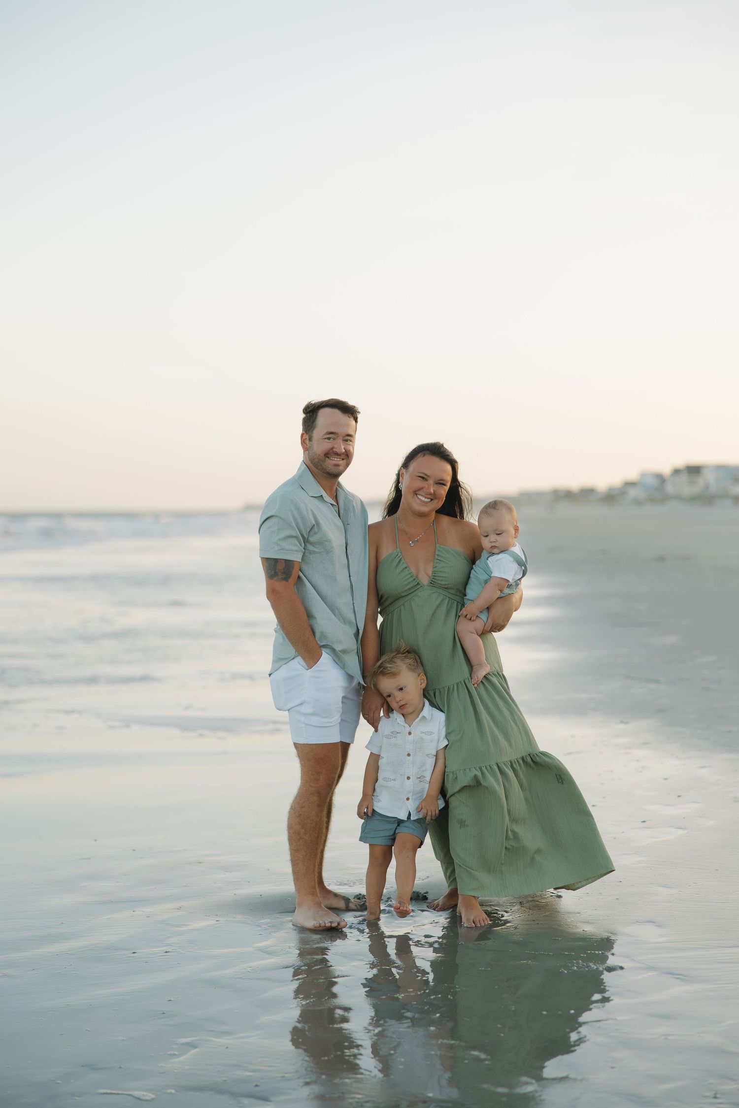 Family of four posing on a beach at sunset, parents in green and white outfits with two young children by the ocean.