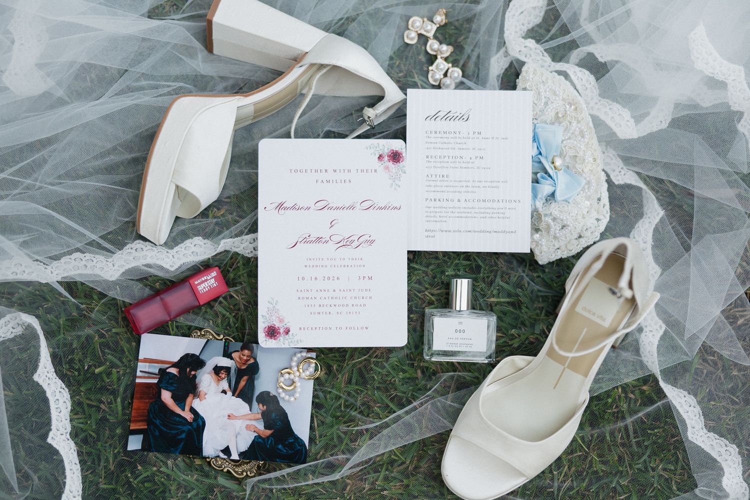 Flat lay of wedding details with ivory heels, invitation suite, perfume bottle, red lipstick, photo and lace veil on green grass.