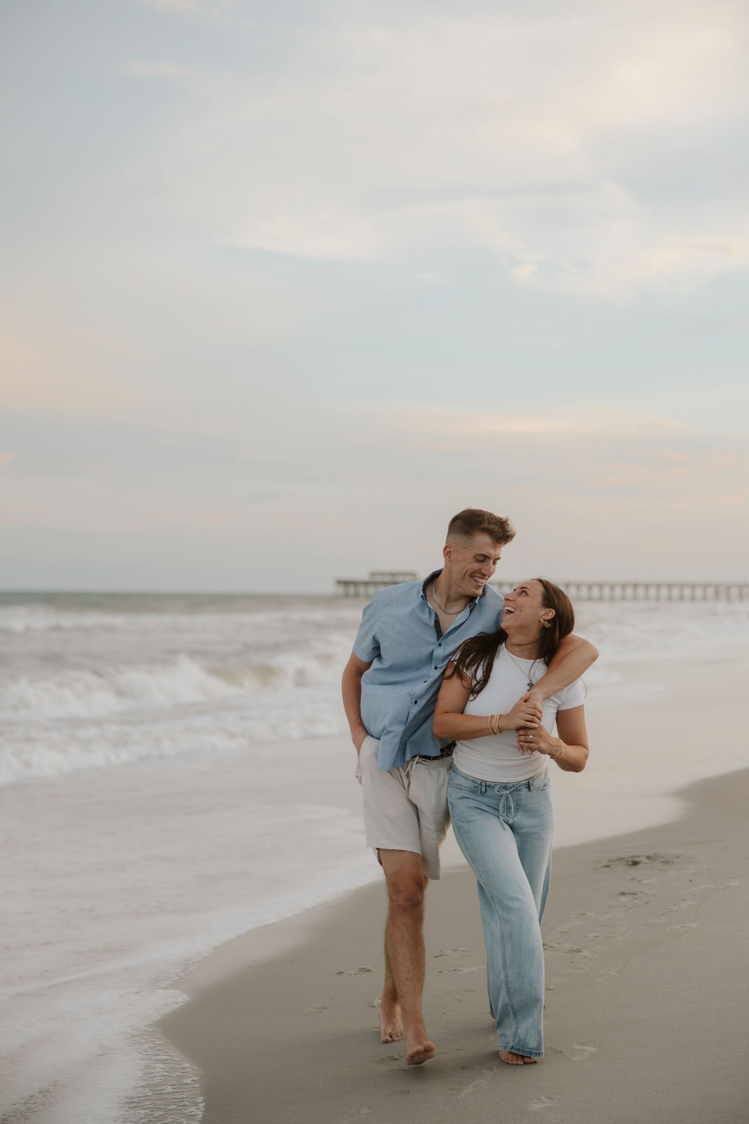 A happy couple embraces while walking on a sandy beach near a pier at sunset.
