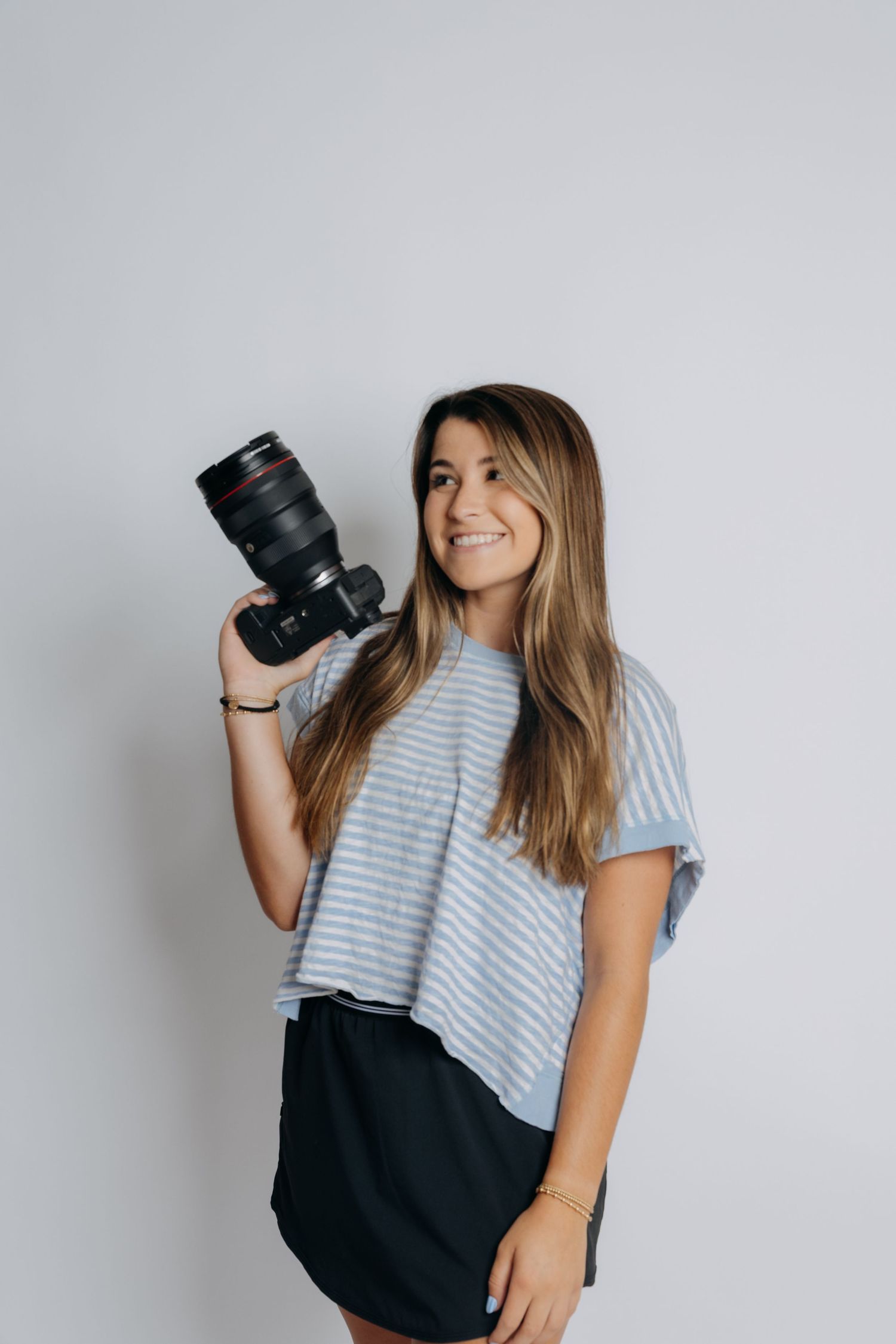 A smiling woman in a striped tee and black skirt holds a Canon DSLR camera against a white background.