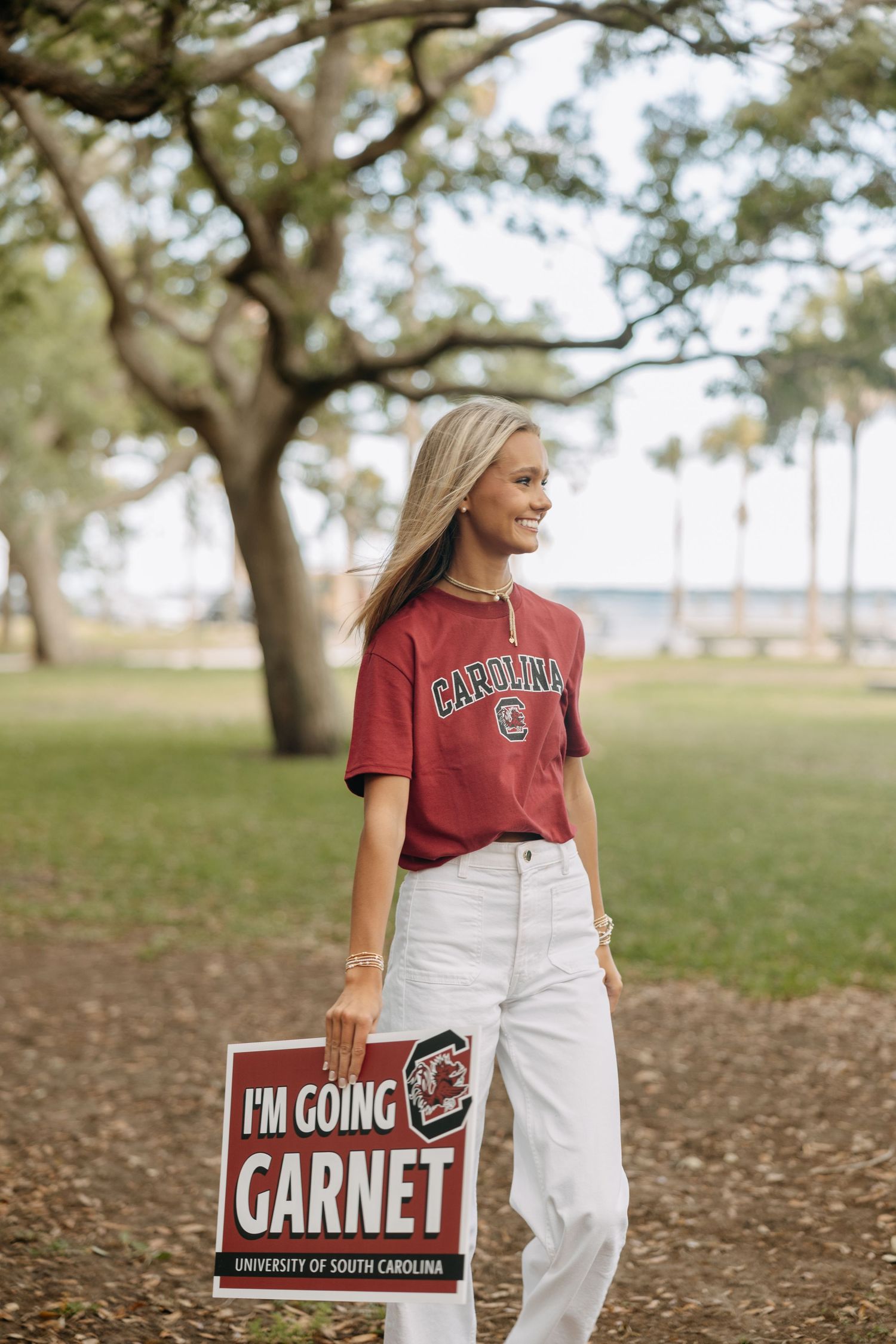 Smiling blonde student holding University of South Carolina Garnet sign in a park setting.