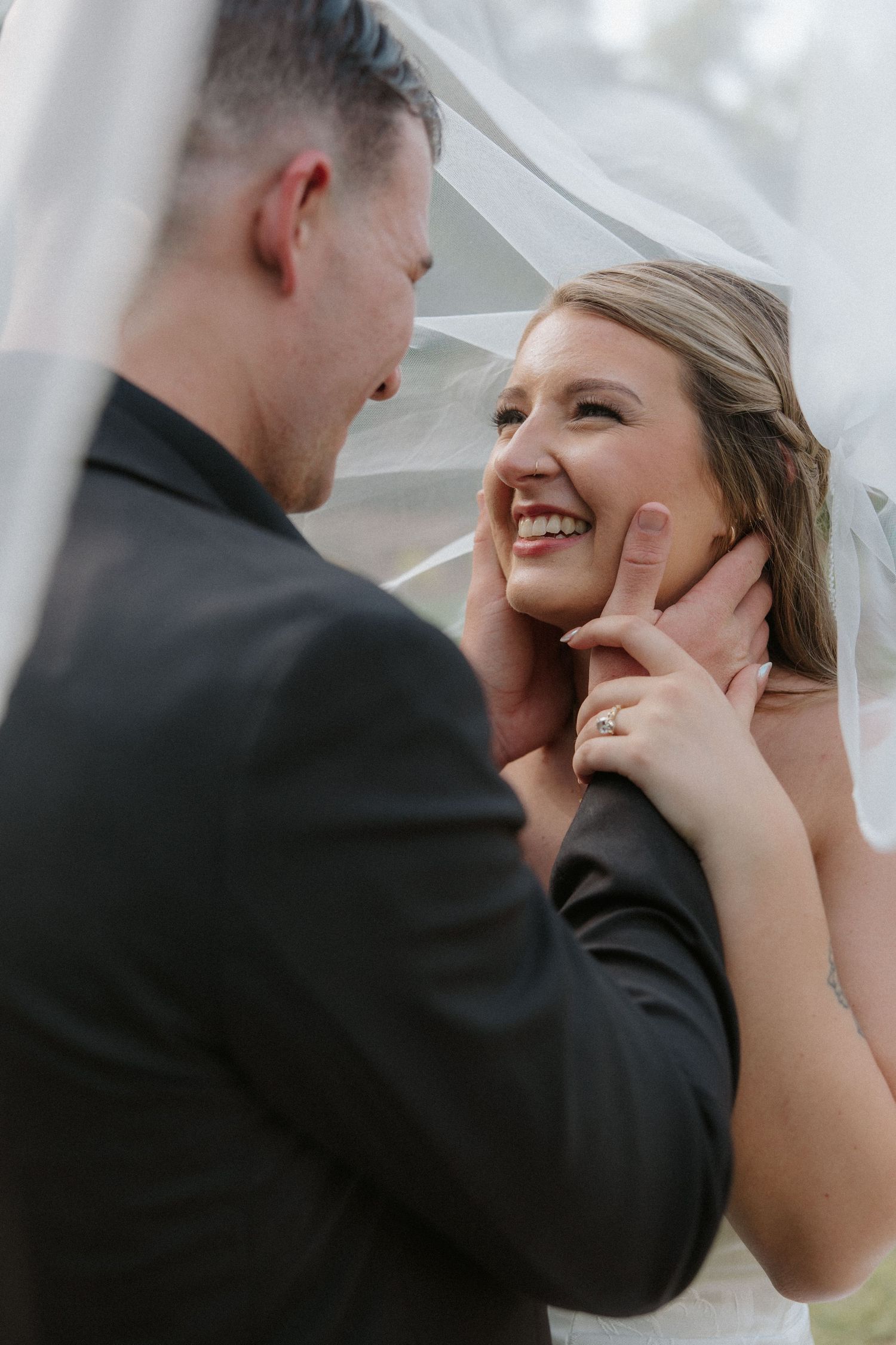 Laughing bride gazing at groom under wedding veil, showcasing diamond engagement ring.