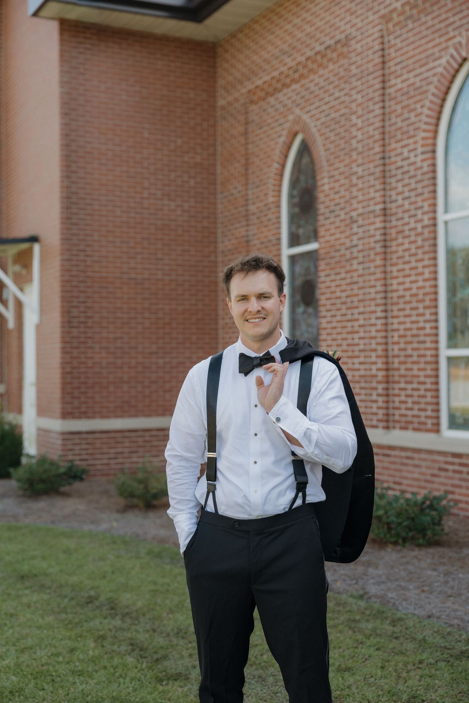 Smiling groom in a white dress shirt, black bow tie and suspenders holds his tuxedo jacket over his shoulder outside a brick church.