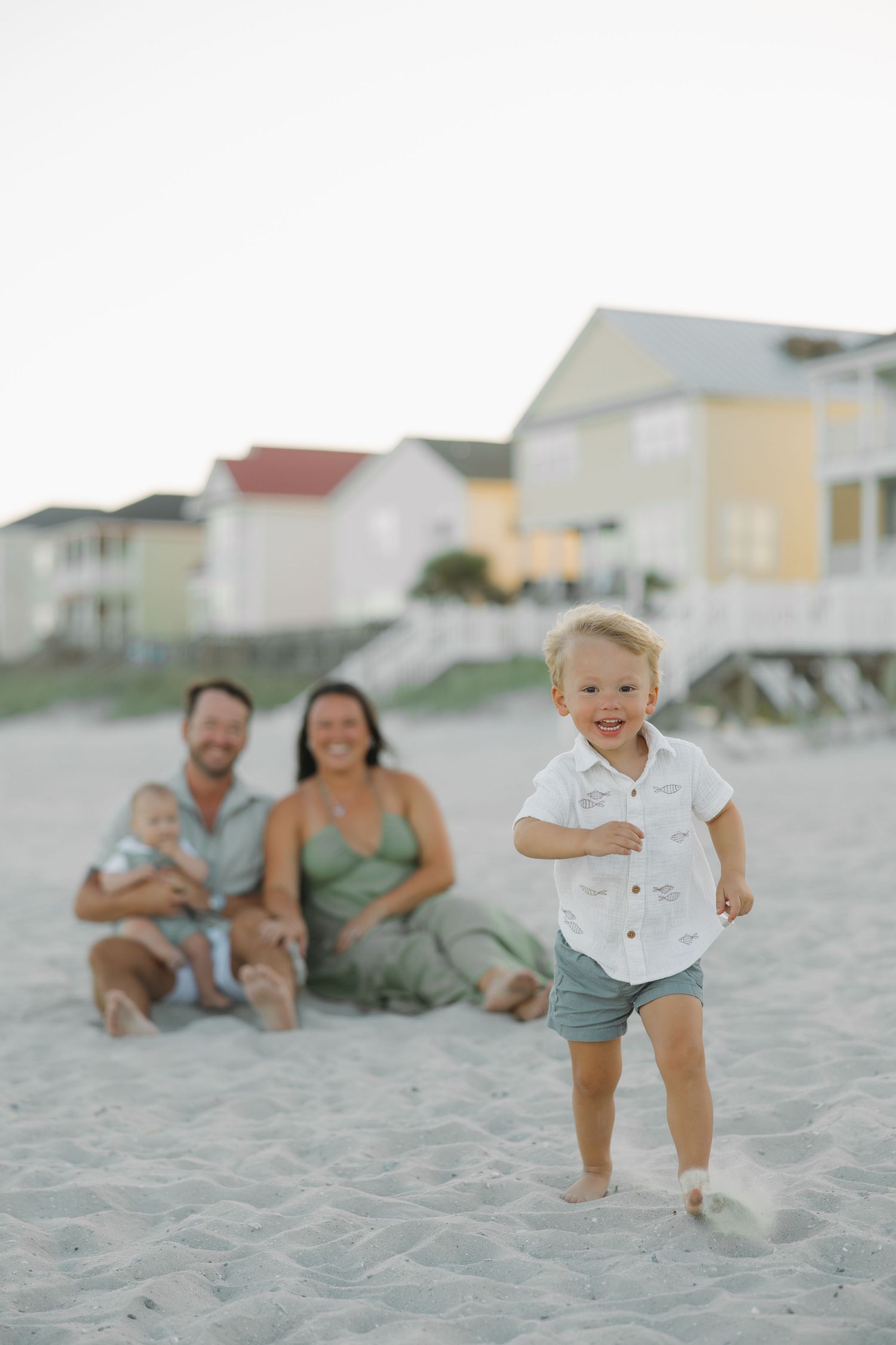 A toddler runs on a sandy beach while his family sits behind him near colorful beach houses at dusk.